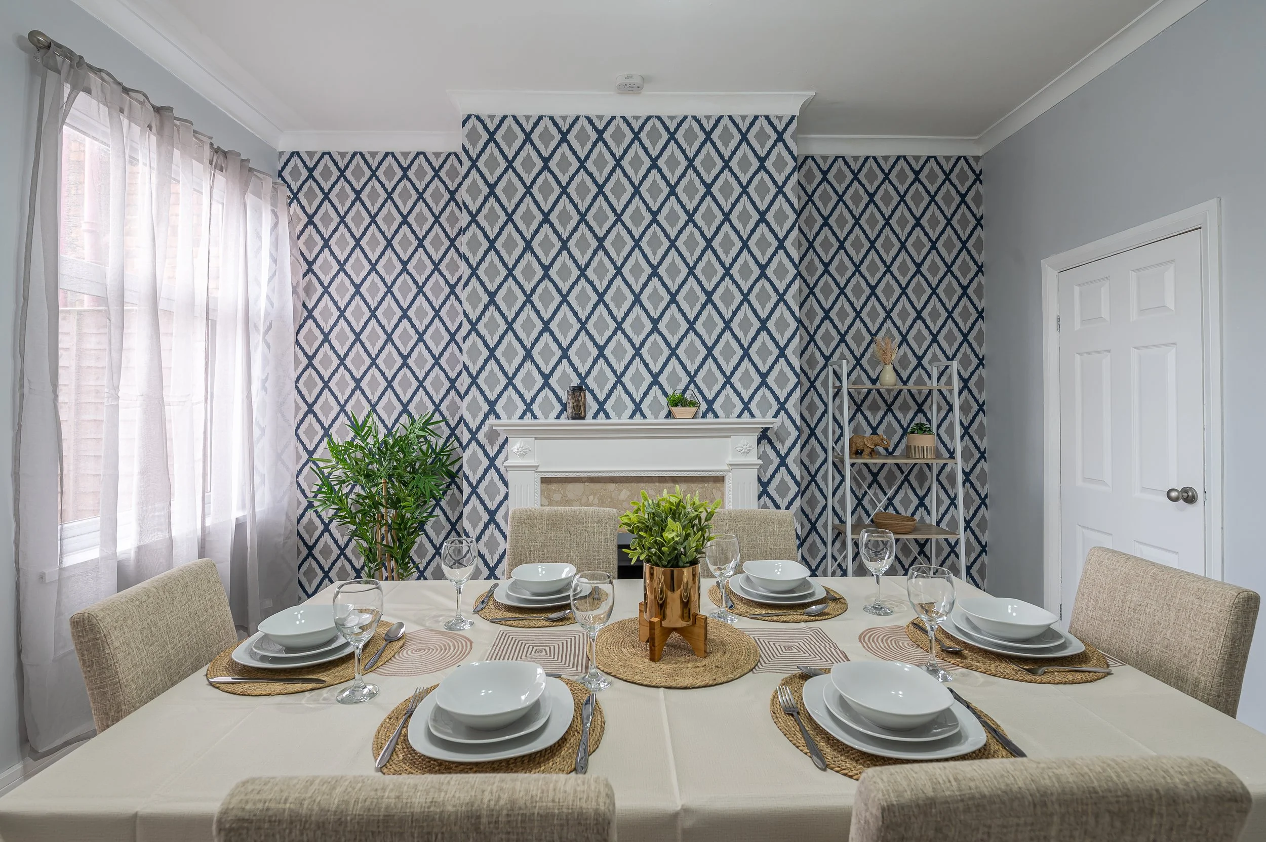 A dining room set with six place settings, including white plates, silverware, wine glasses, and woven placemats on a beige tablecloth. In the center, there are two wooden vases with greenery and a small decorative object. The room features a pattern