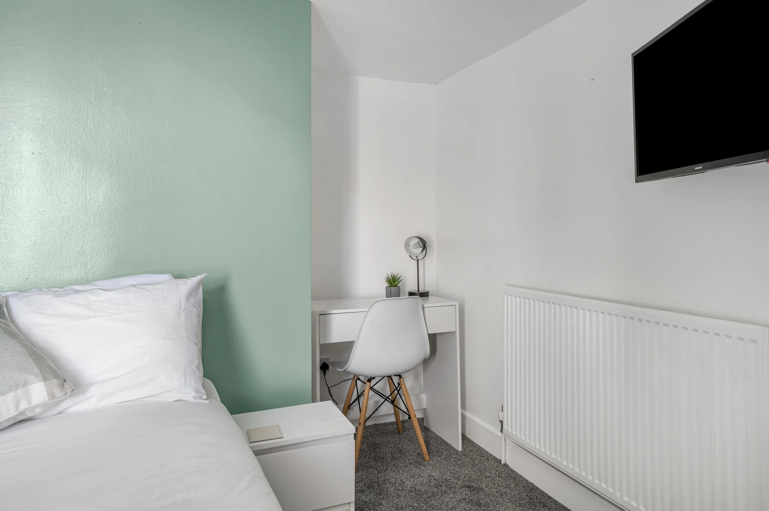 A bedroom corner with a white bed, a white nightstand, a small desk with a white chair, a small potted plant, a wall-mounted TV, a wall with a green accent color, and a gray carpet.