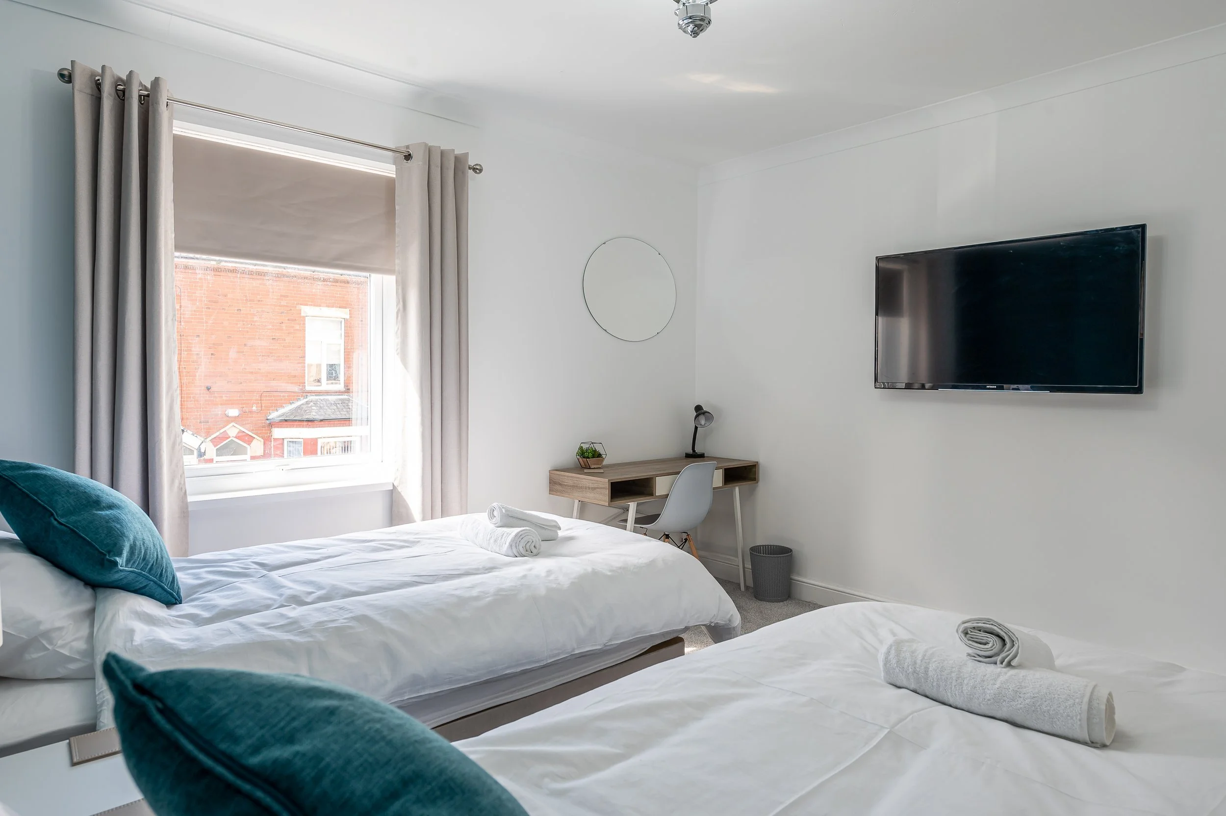 A bright bedroom with white walls, a window with beige curtains, a wall-mounted flat-screen TV, a small desk with a lamp and decorative plant, and 2 beds with turquoise pillows and rolled towels.