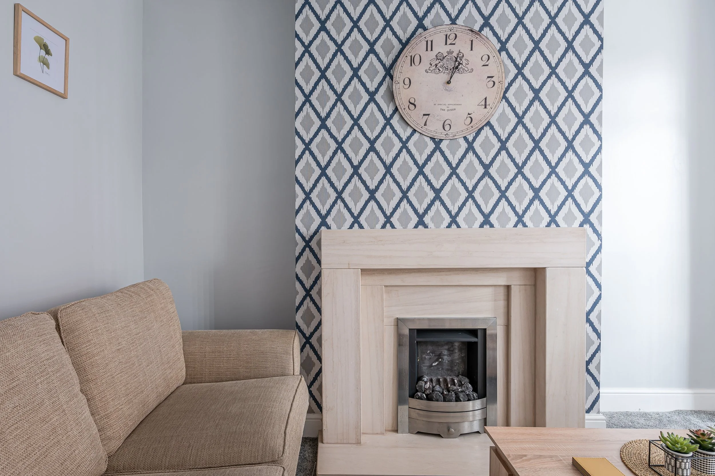 Living room with beige sofa on the left, a wall with geometric blue and gray wallpaper behind a fireplace, a large round clock above the fireplace, and a small wooden table with decorative items in the foreground.