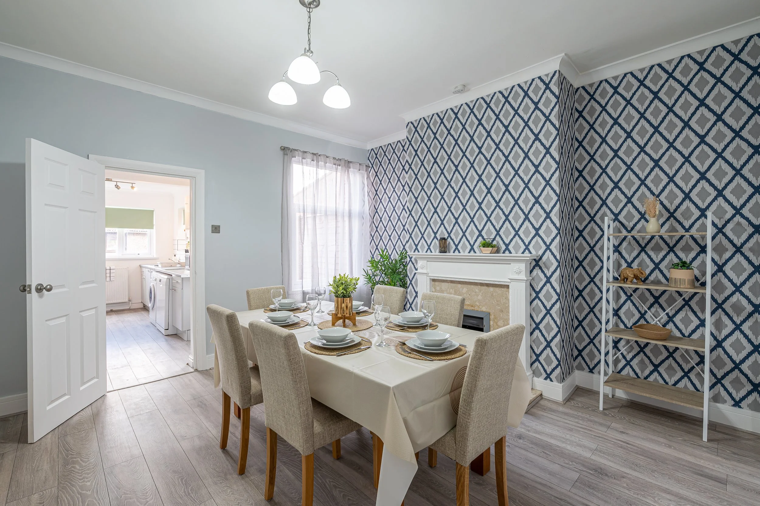 A dining room with a dining table set with six place settings, overlooking a living space with a blue and gray patterned accent wall, a white fireplace, and a shelf with decorative items. A window with sheer curtains and a door leading to a kitchen are also visible.