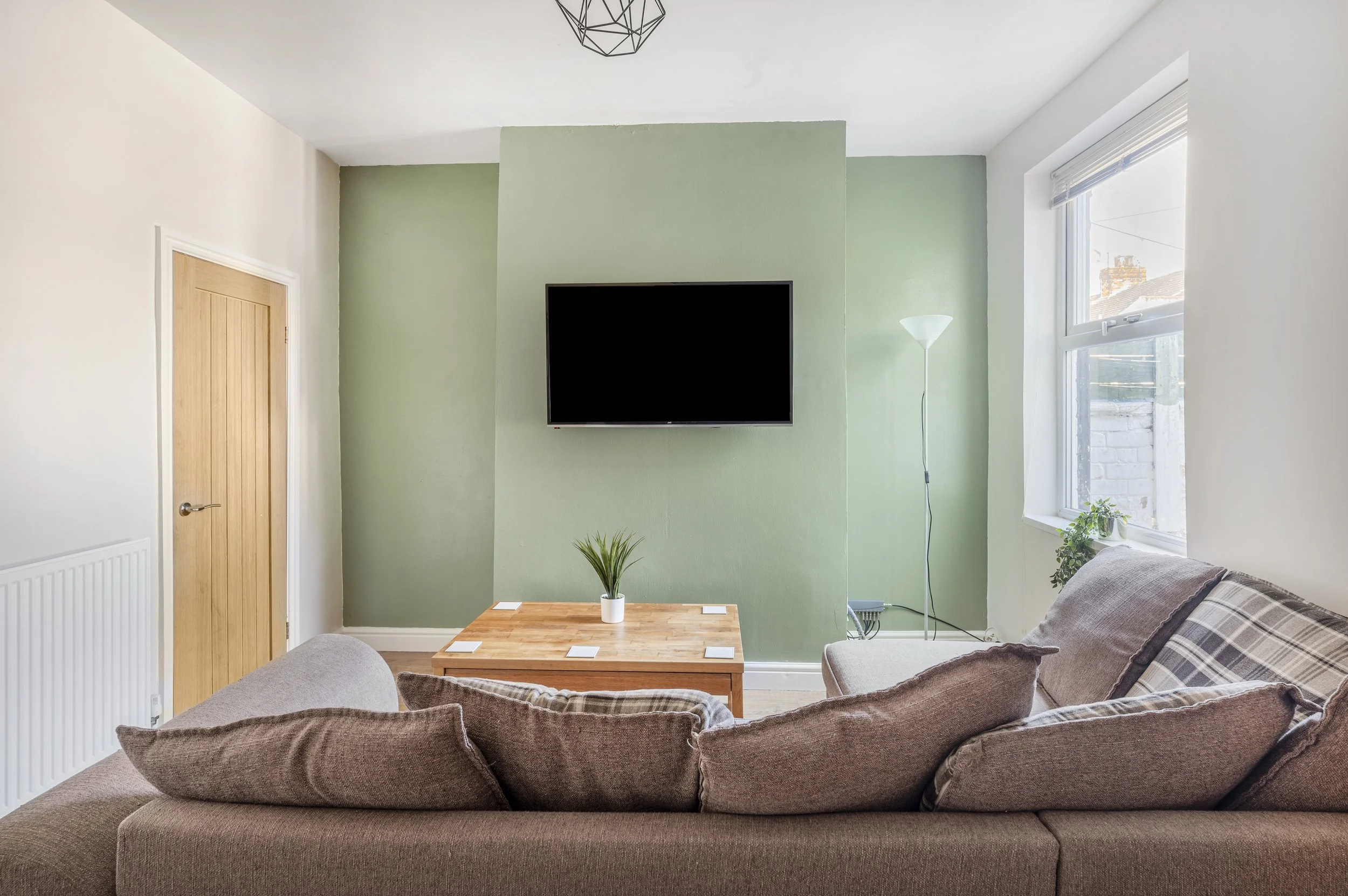Living room with a brown sofa, a wooden coffee table, a green accent wall, a flat-screen TV, a floor lamp, a large window with blinds, and a plant on the windowsill.