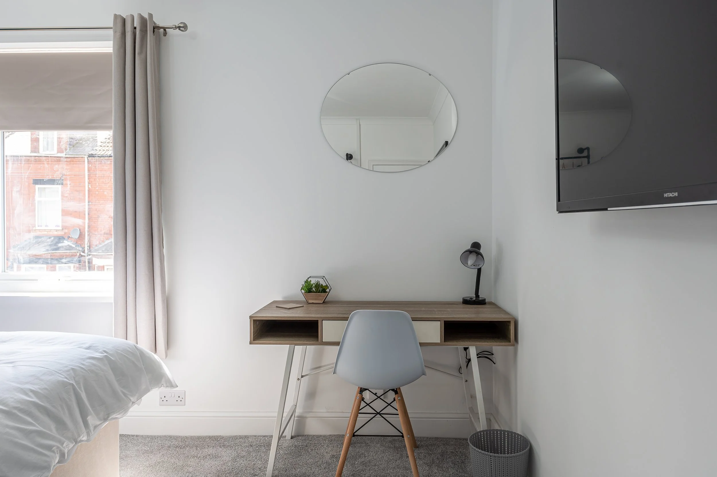 A bedroom with a window covered by light gray curtains, a round mirror on the white wall, and a small wooden desk with a potted plant, a desk lamp, and a chair. A television is mounted on the wall. The room has a neutral color scheme with gray carpet