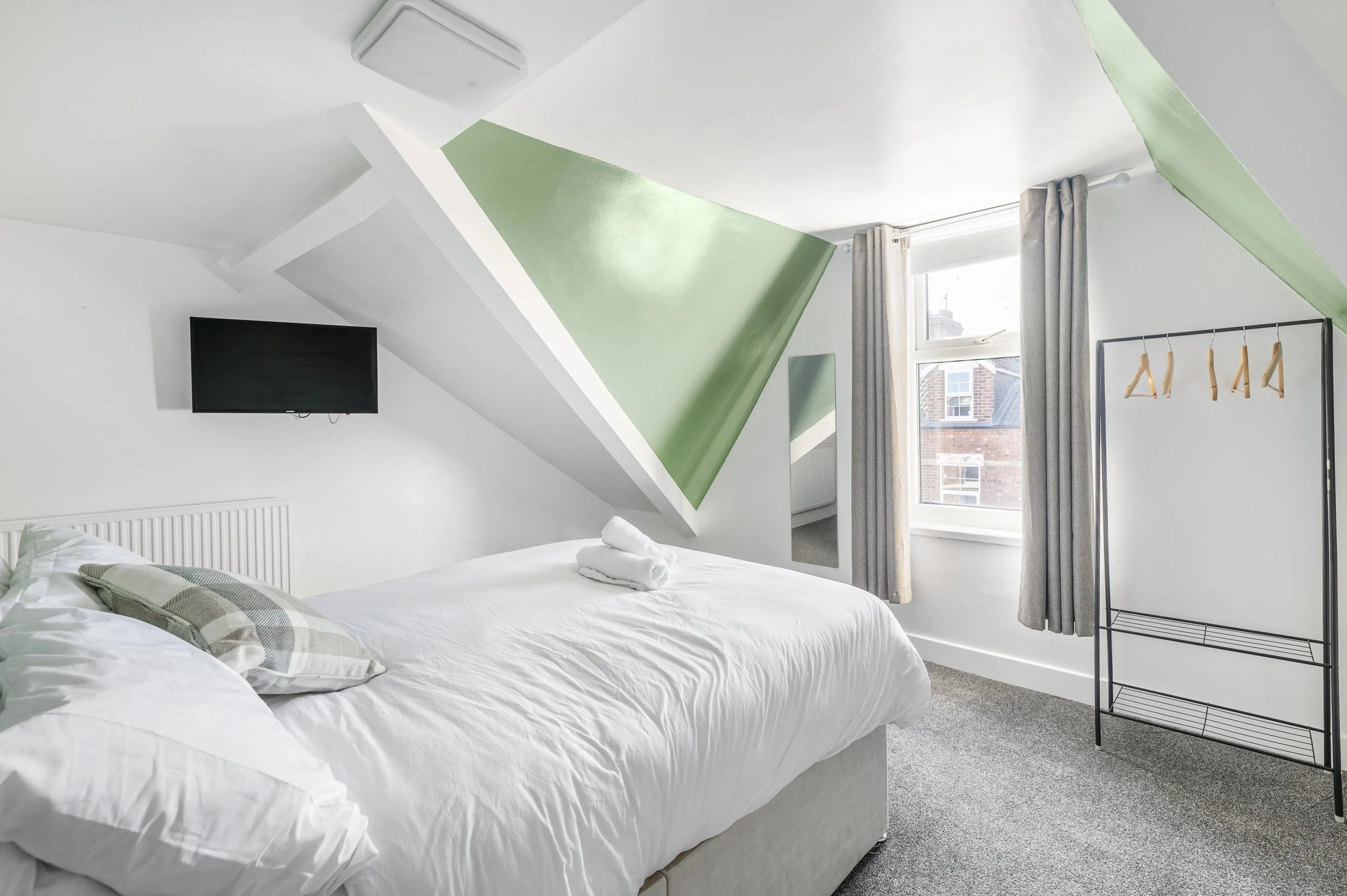 A bedroom with white walls and a sloped ceiling painted green. There is a large window with gray curtains, a flat-screen TV mounted on the wall, a bed with white linens and a plaid pillow, and a black metal clothing rack with wooden hangers. Towels a