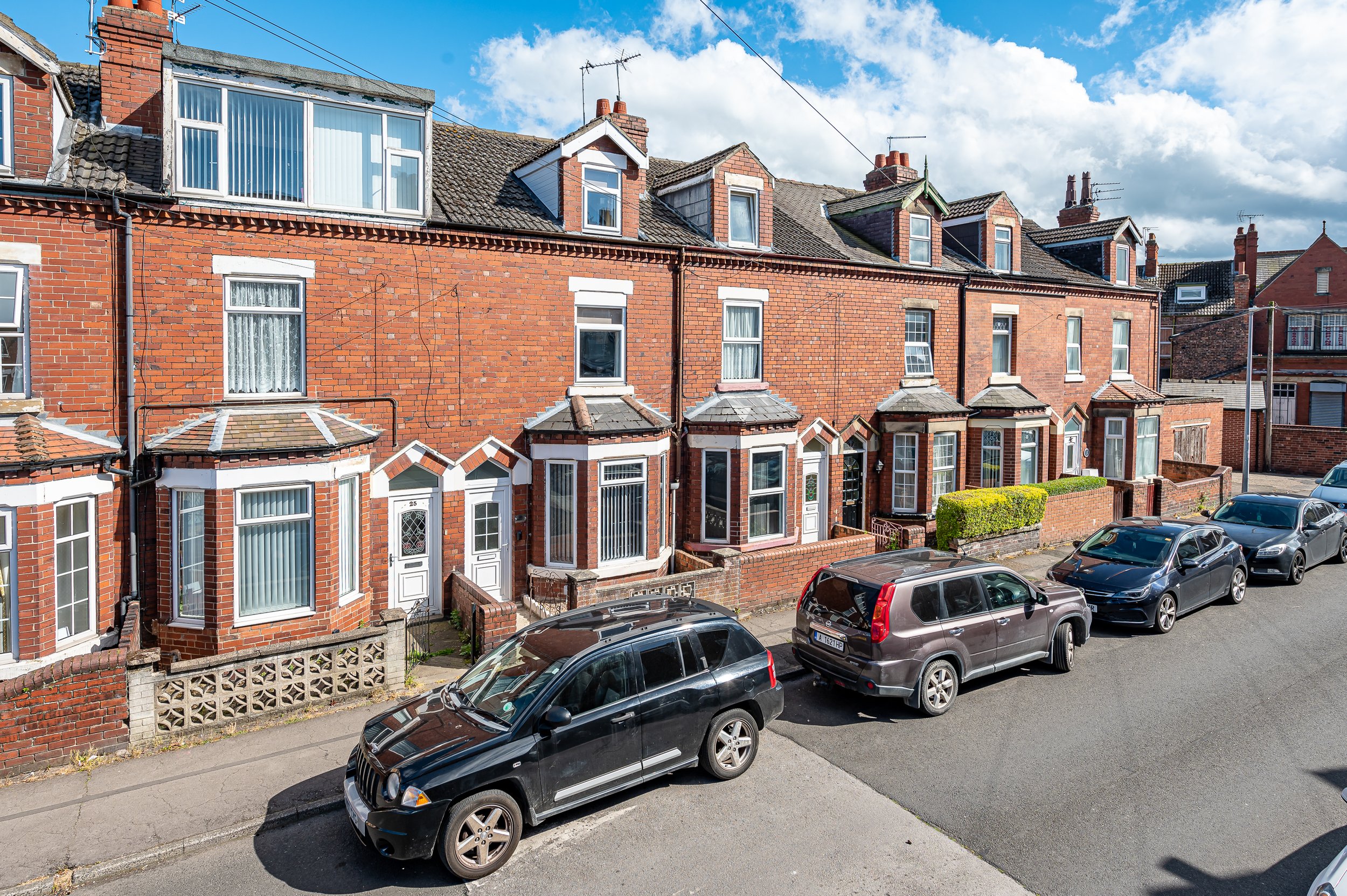 Row of brick houses with variously shaped windows and roofs, parked cars along the street in front, under a blue sky with clouds.