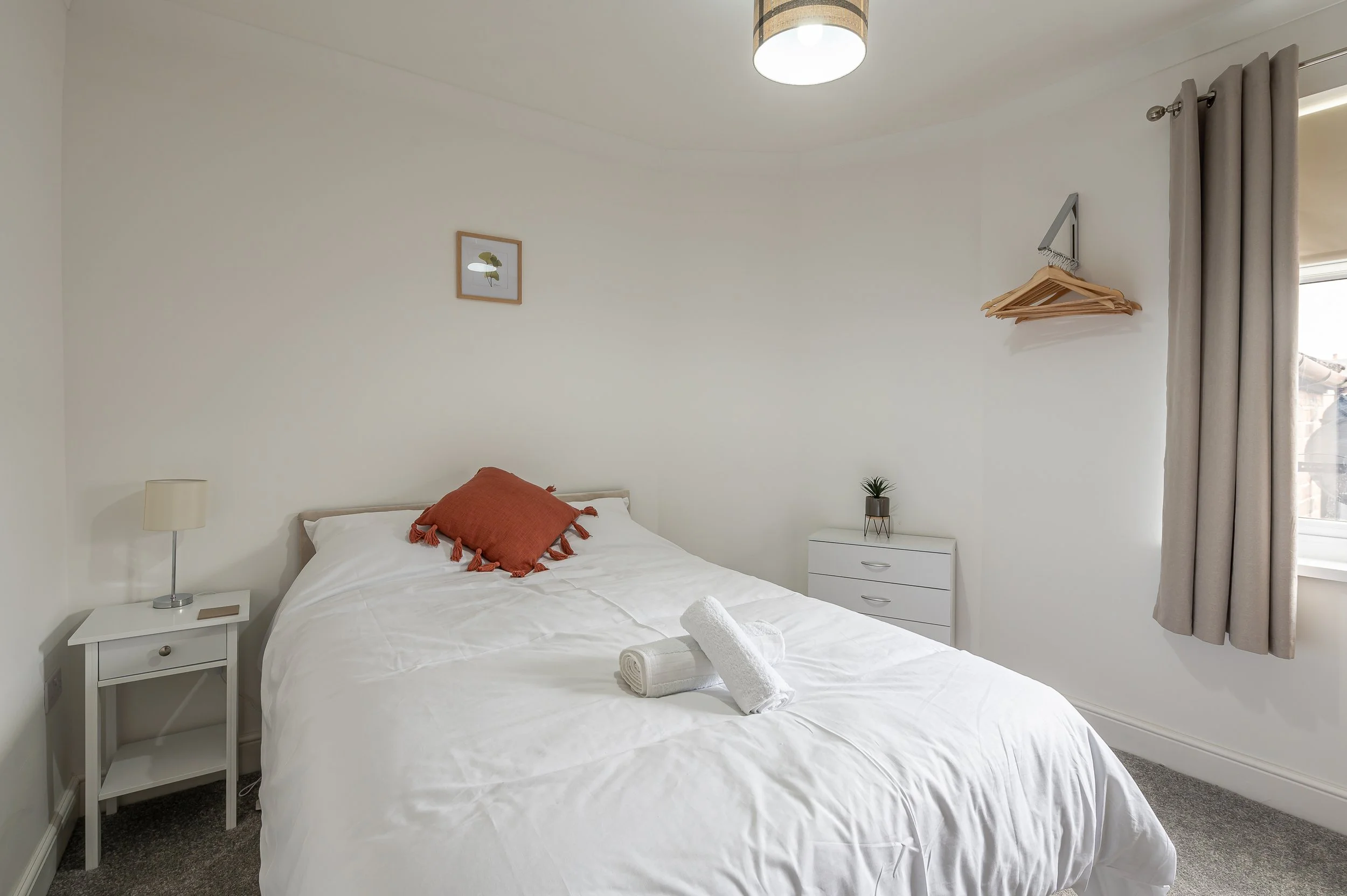 A neatly made bed with white bedding and a rust-colored throw pillow in a minimalist bedroom. There is a bedside table with a lamp and framed artwork of a leaf on the wall. A small white dresser with a potted plant is beside the window, which has bei