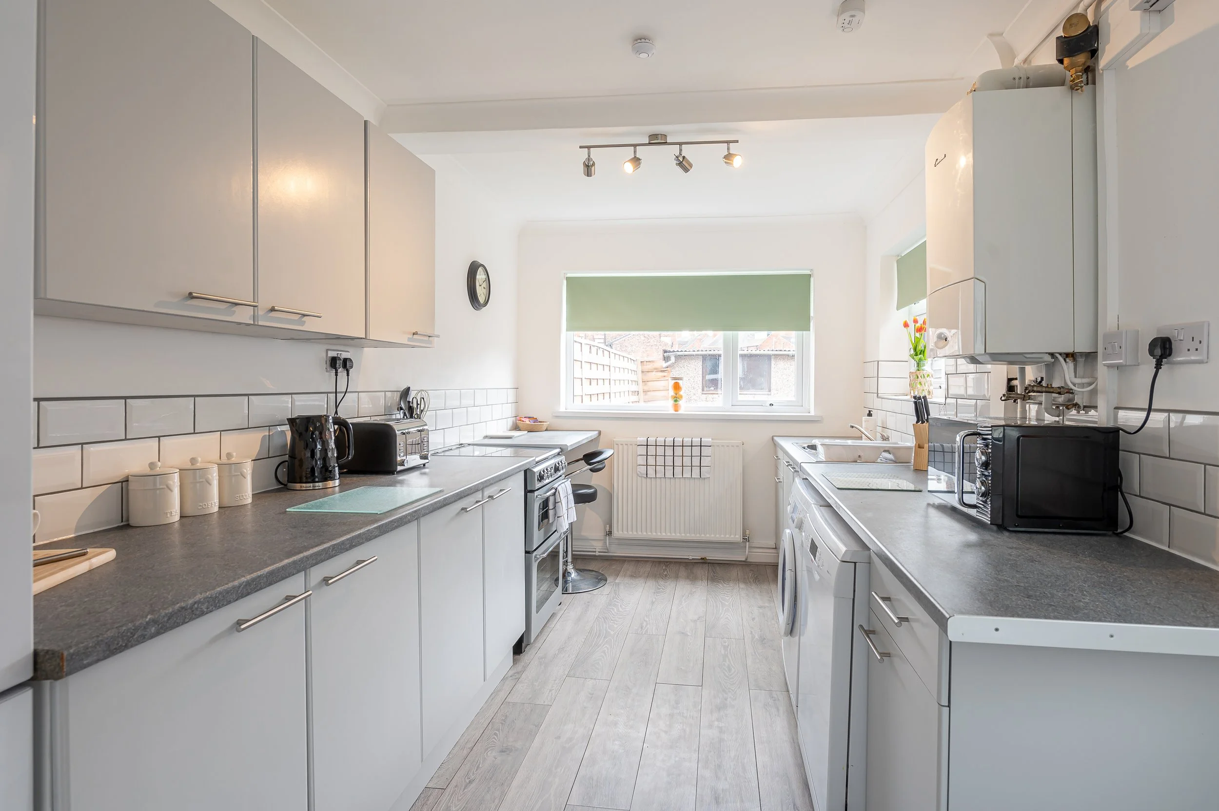 A kitchen with white cabinets, gray countertops, and a window with a green roller blind. The kitchen has appliances including a microwave, toaster, kettle, and a washing machine. There are decorative canisters on the counter and a vase with tulips ne