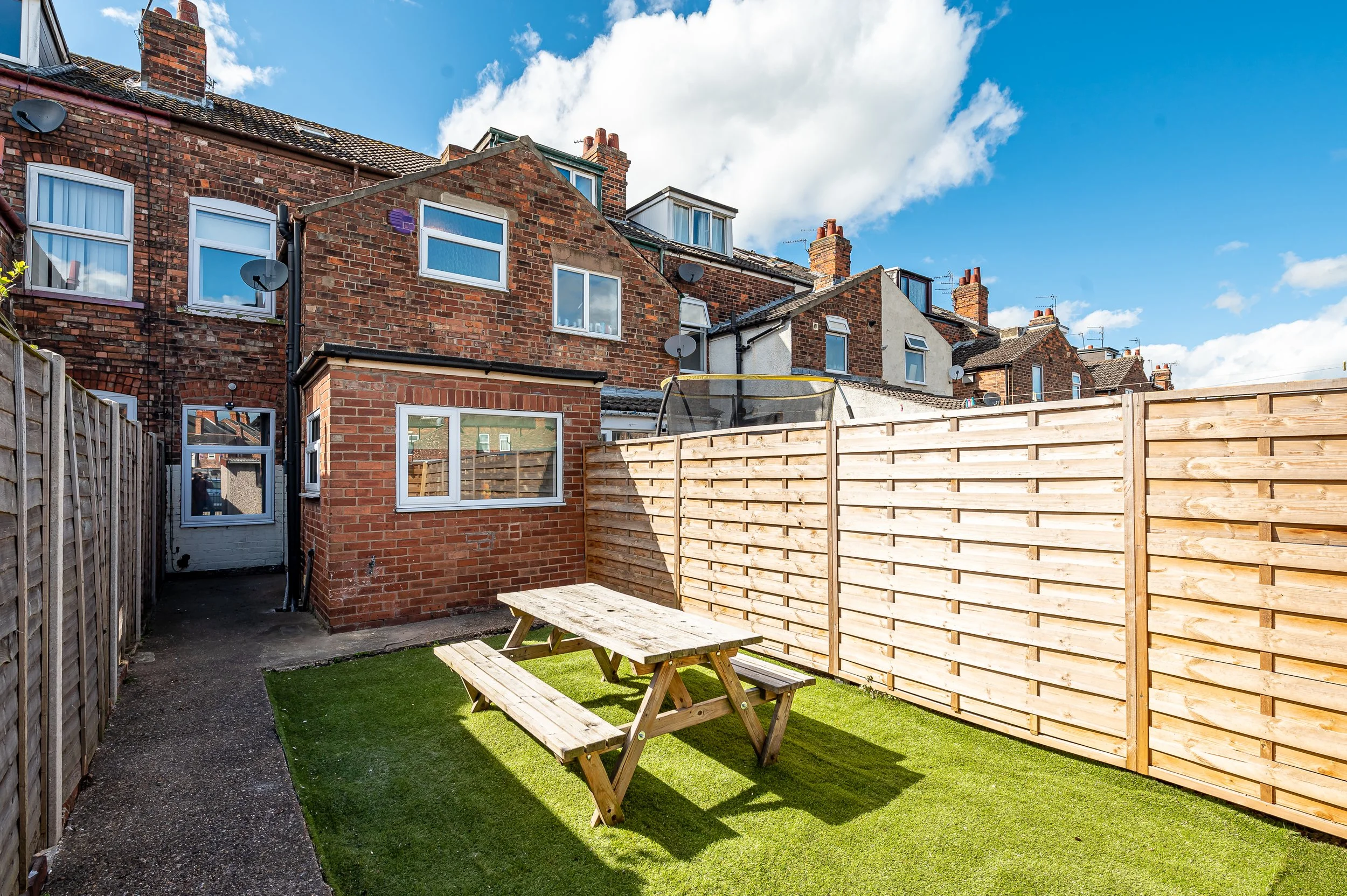 A small backyard with artificial grass, a wooden picnic table, and a tall wooden fence. The background features brick houses with multiple windows and chimneys, satellite dishes, and a partly cloudy sky.