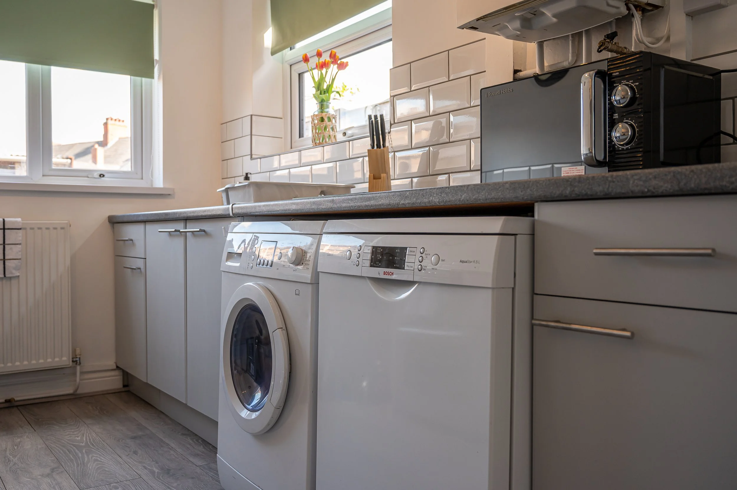 A kitchen during daytime with a white washing machine, a kitchen counter with a knife holder, a microwave, and a small window with a vase of tulips on the sill, white tiled backsplash, and a radiator on the wall.