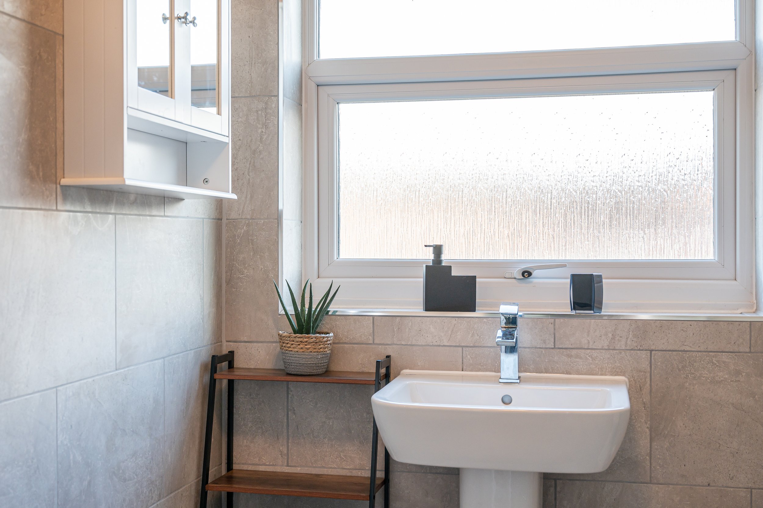 Bathroom with a large window, a white bathtub, a faucet, a potted plant on a small wooden table, a mirror cabinet, and beige tiled walls.