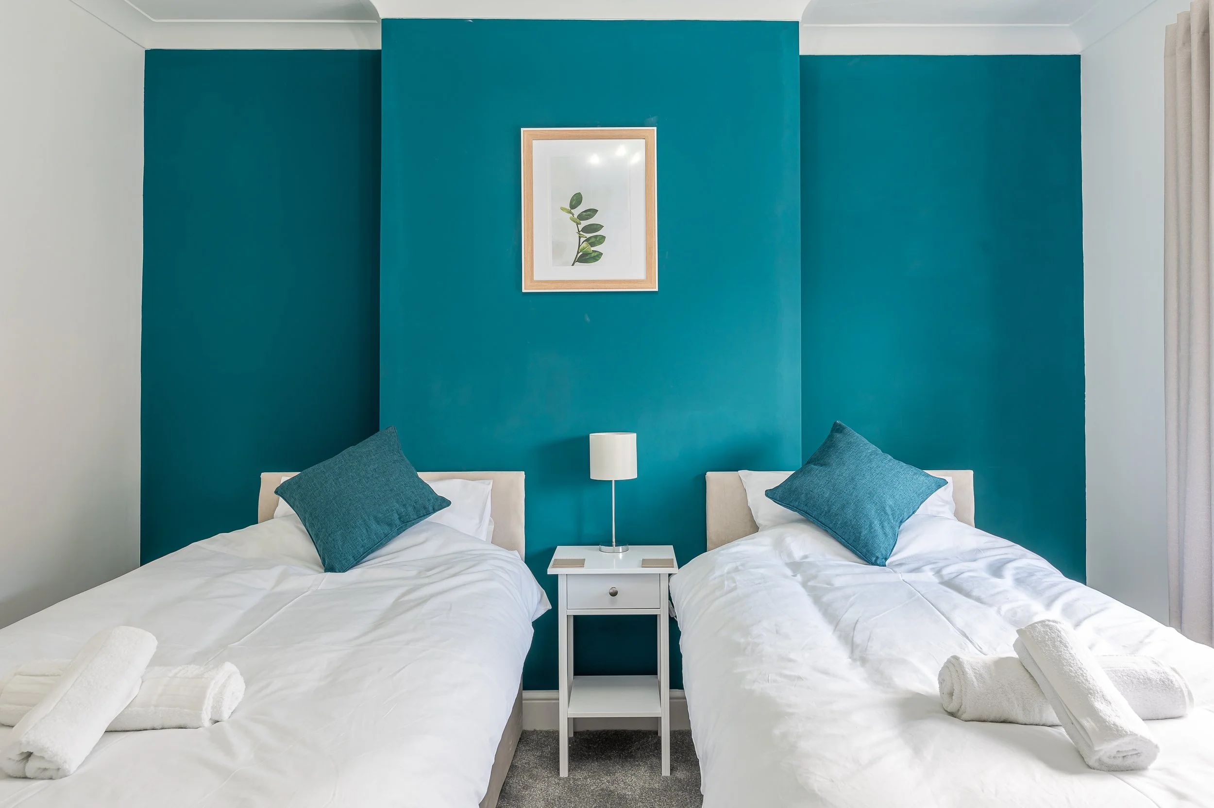 Two beds with white linens, each with two dark blue pillows and rolled white towels on top, are placed beside each other separated by a small white nightstand. The nightstand holds a white lamp and a small book. The beds are positioned against a teal