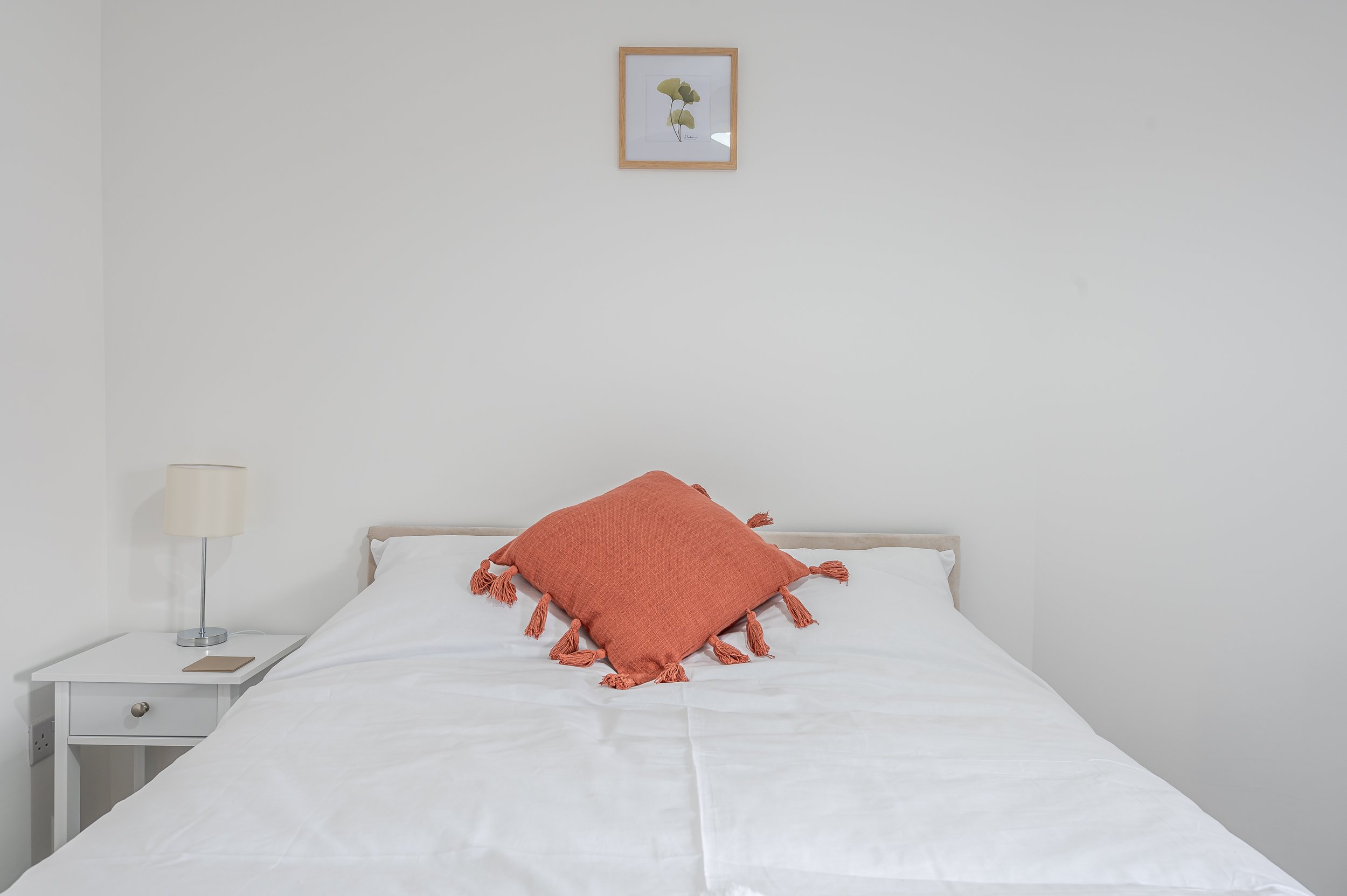 A neatly made bed with white sheets and a large orange pillow with tassels. A white nightstand with a lamp and a smartphone. White walls with a framed botanical print hanging above the bed. Minimalist decor in a modern bedroom.