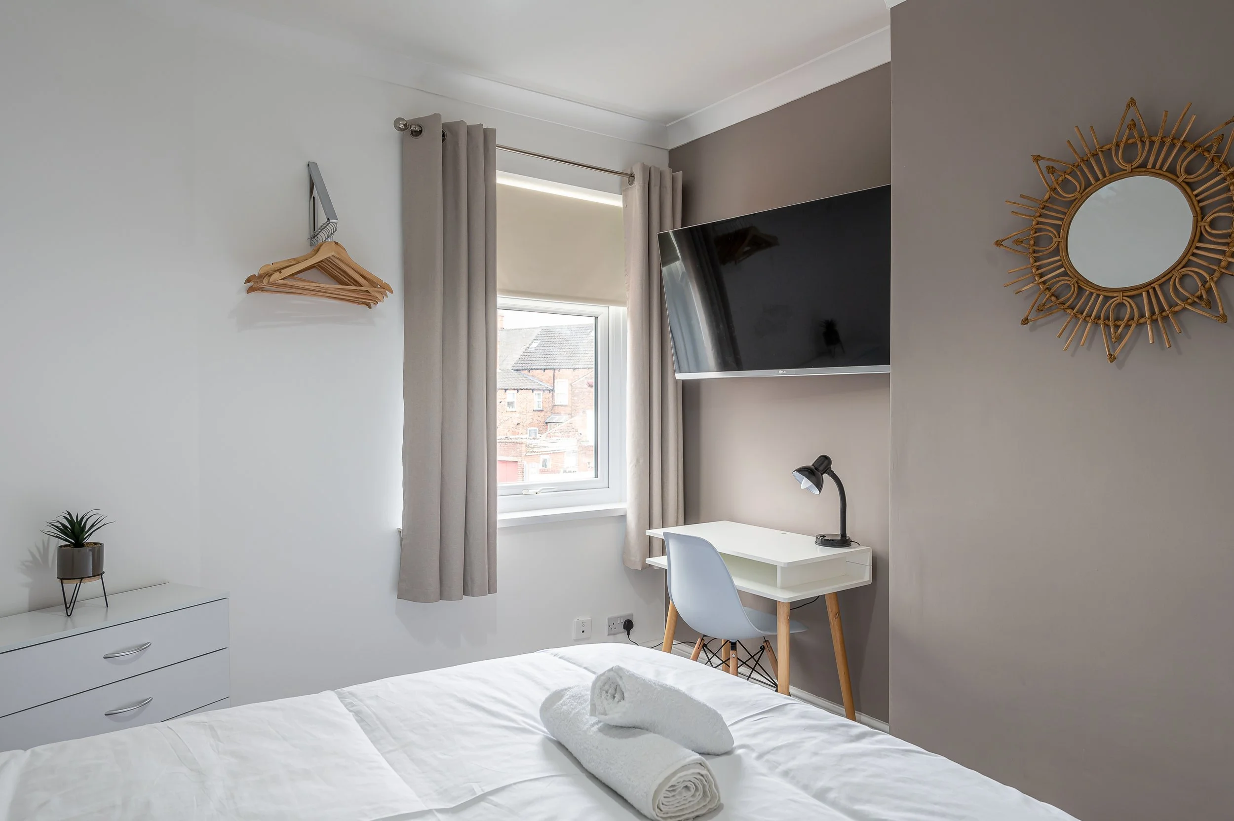 A tidy bedroom with white walls, a window with beige curtains, a wall-mounted TV, a white desk with a chair, a black desk lamp, a mirror with a decorative rattan frame, a white dresser with a small potted plant, and neatly folded towels on the bed.