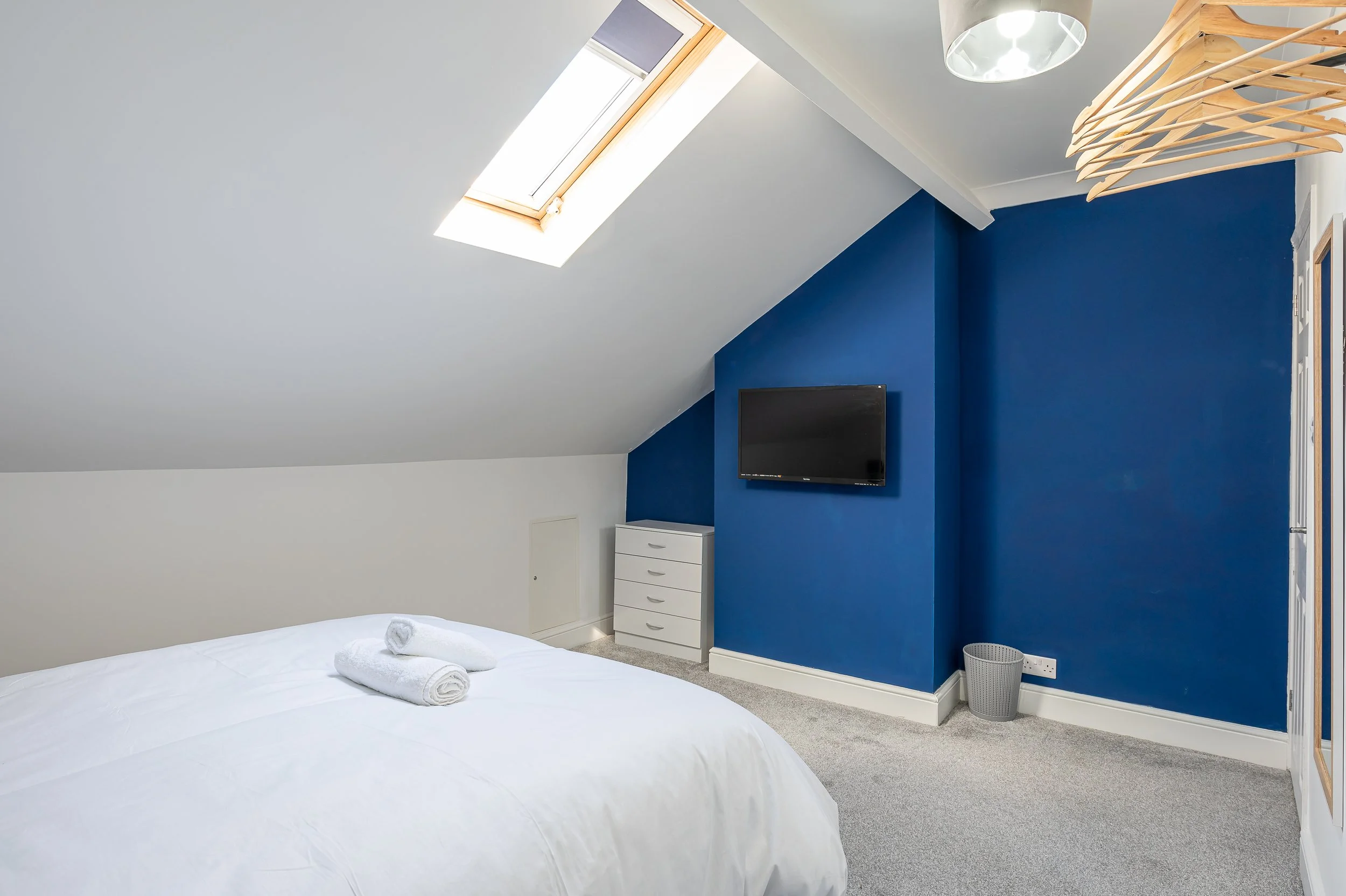 A bedroom with a sloped ceiling, white walls, and a blue accent wall. There's a bed with white bedding and rolled towels, a small white dresser, a wall-mounted TV, a ceiling light, a laundry basket, a window, and a wooden coat hanger rod.