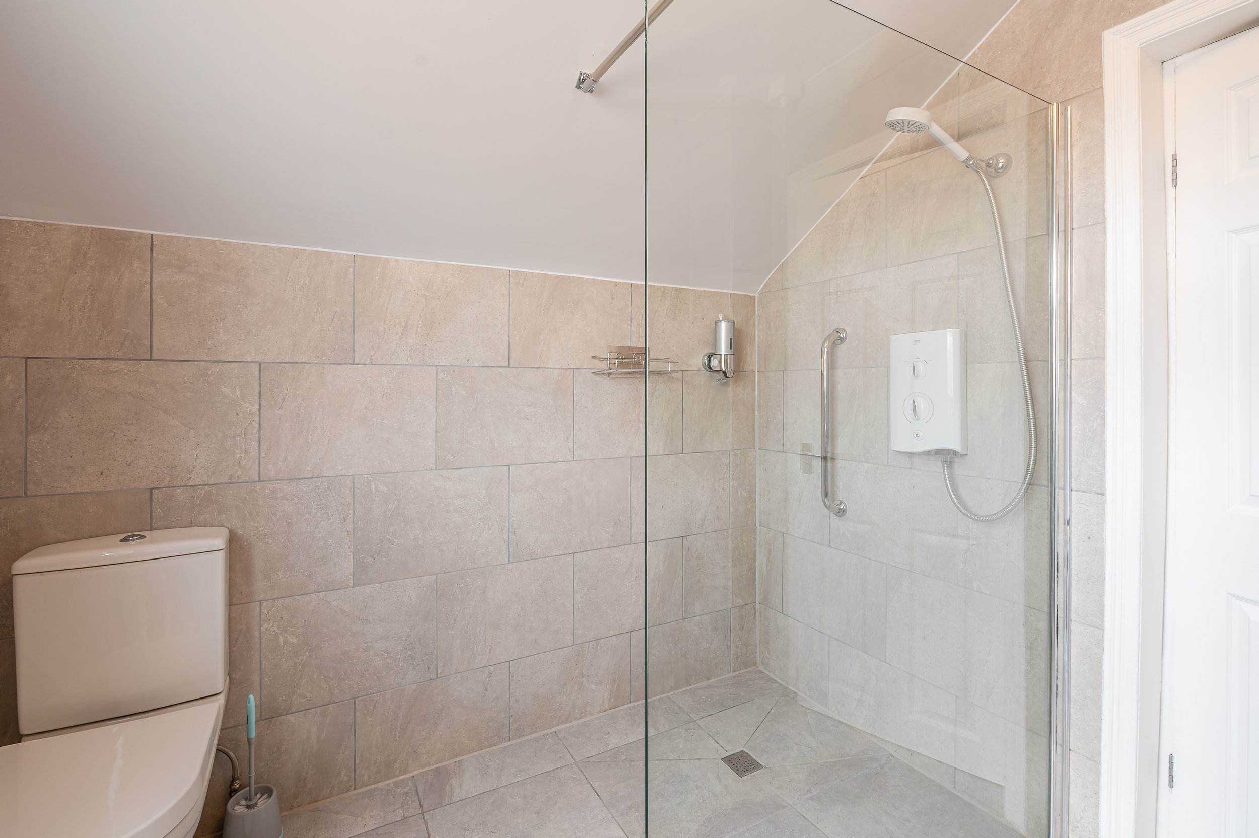 Bathroom with beige tiled walls and floor, a glass shower enclosure with a showerhead and controls, a white toilet, a wall-mounted soap dispenser, a shelf, a grab bar, and a floor drain.
