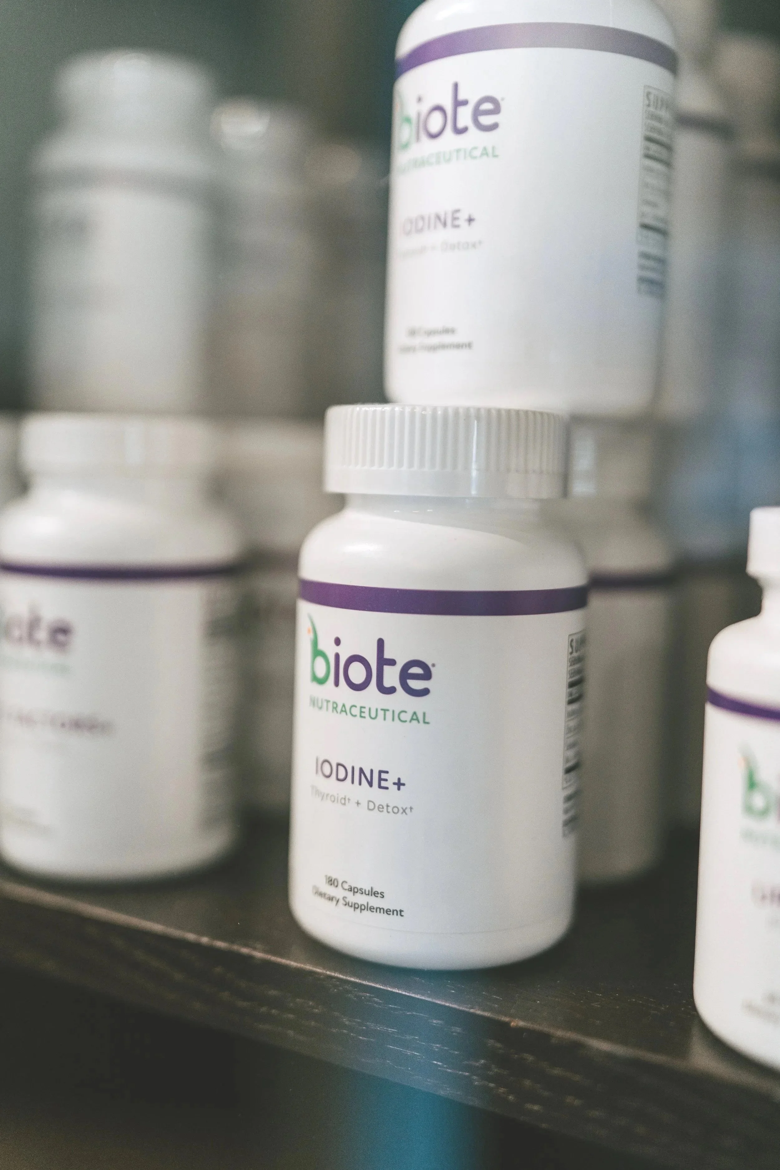 A bottle of Biote NutraCeutical Iodine+ dietary supplement with 140 capsules on a black shelf, with other supplement bottles blurred in the background.