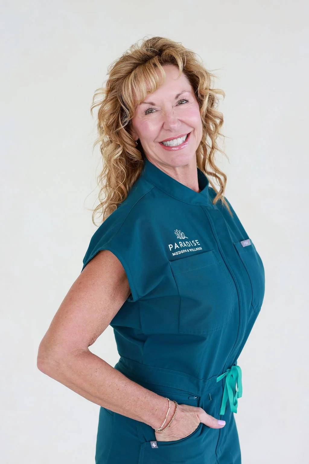 A smiling woman with curly reddish-blonde hair wearing teal medical scrubs with 'Paradise Medspa & Wellness' logo, standing against a plain white background.