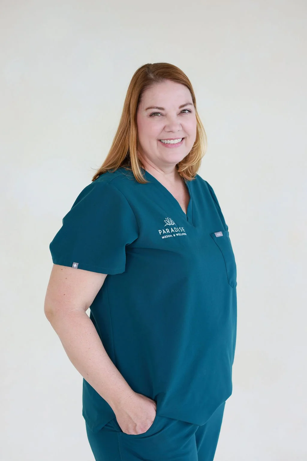 A smiling woman with shoulder-length red hair wearing teal medical scrubs with 'Paradise Massage & Wellness' logo, standing against a plain light-colored background.