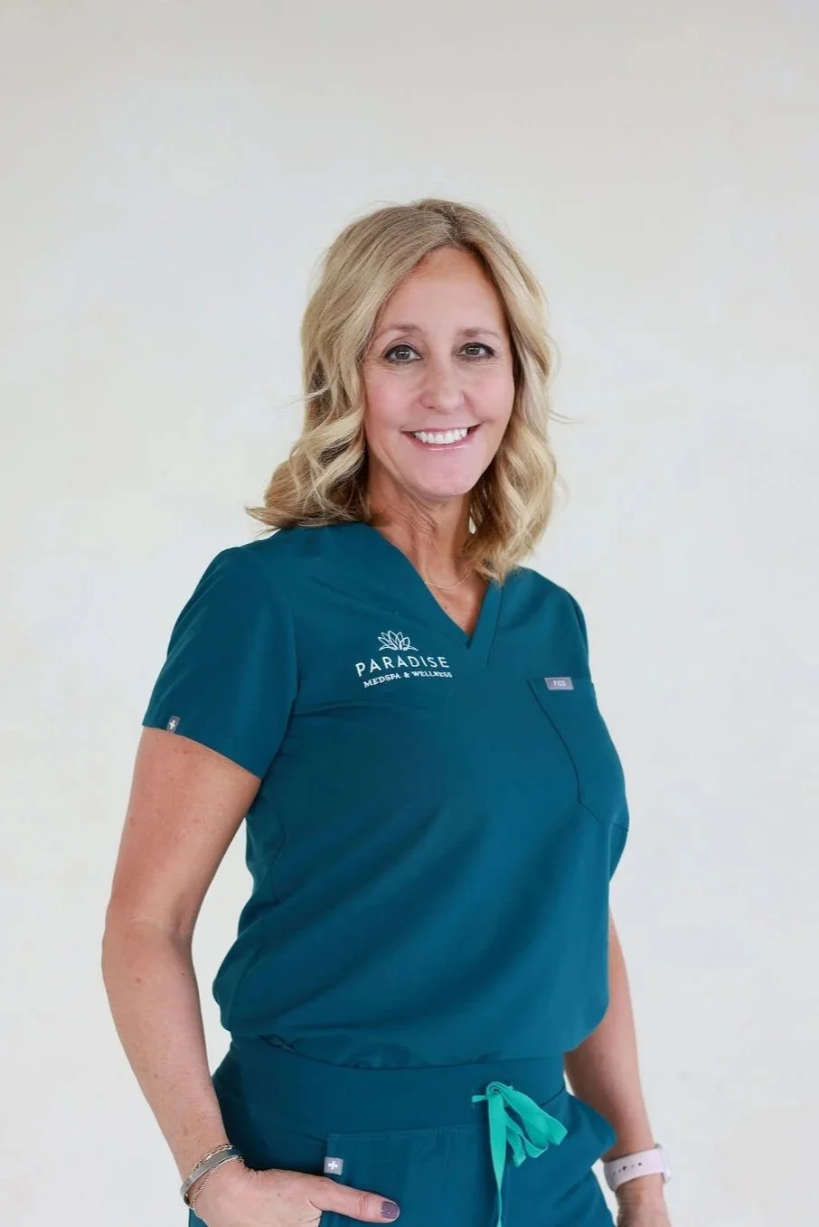A smiling woman in teal medical scrubs with 'Paradise Medspa & Wellness' logo standing against a plain white background.