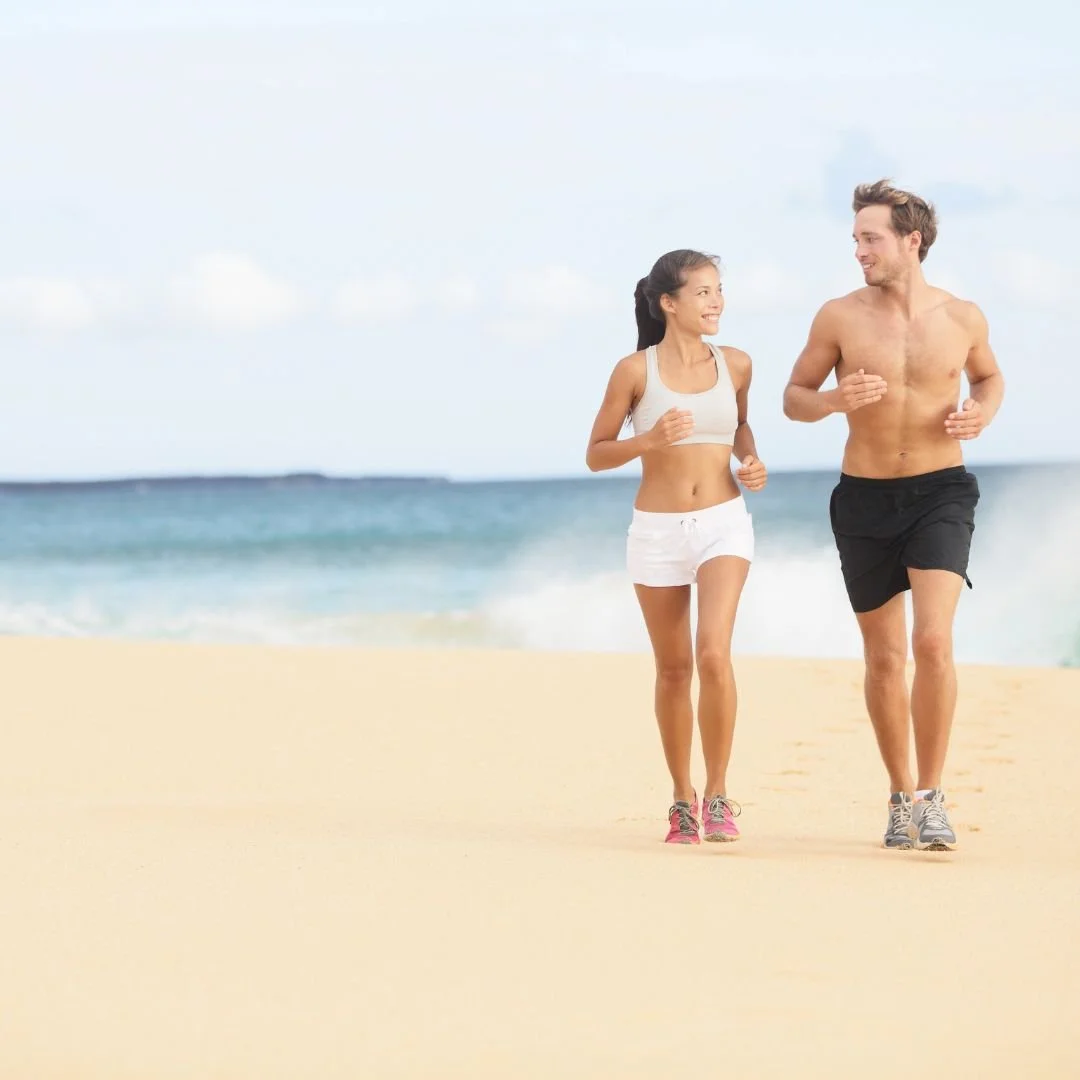 A man and woman jogging on a beach, the man shirtless and wearing black shorts, the woman in a white sports top and shorts, both smiling.