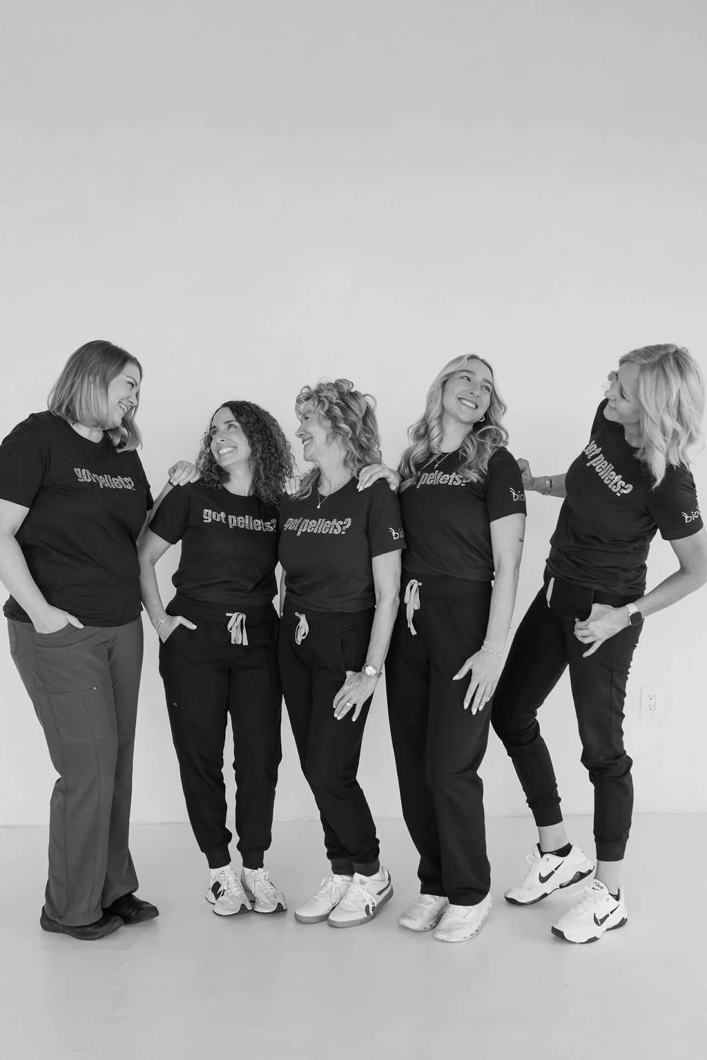 Five women standing together, smiling and looking at each other, wearing matching black T-shirts with the text 'got pellets?' or 'pallets?' in white, in a white room.