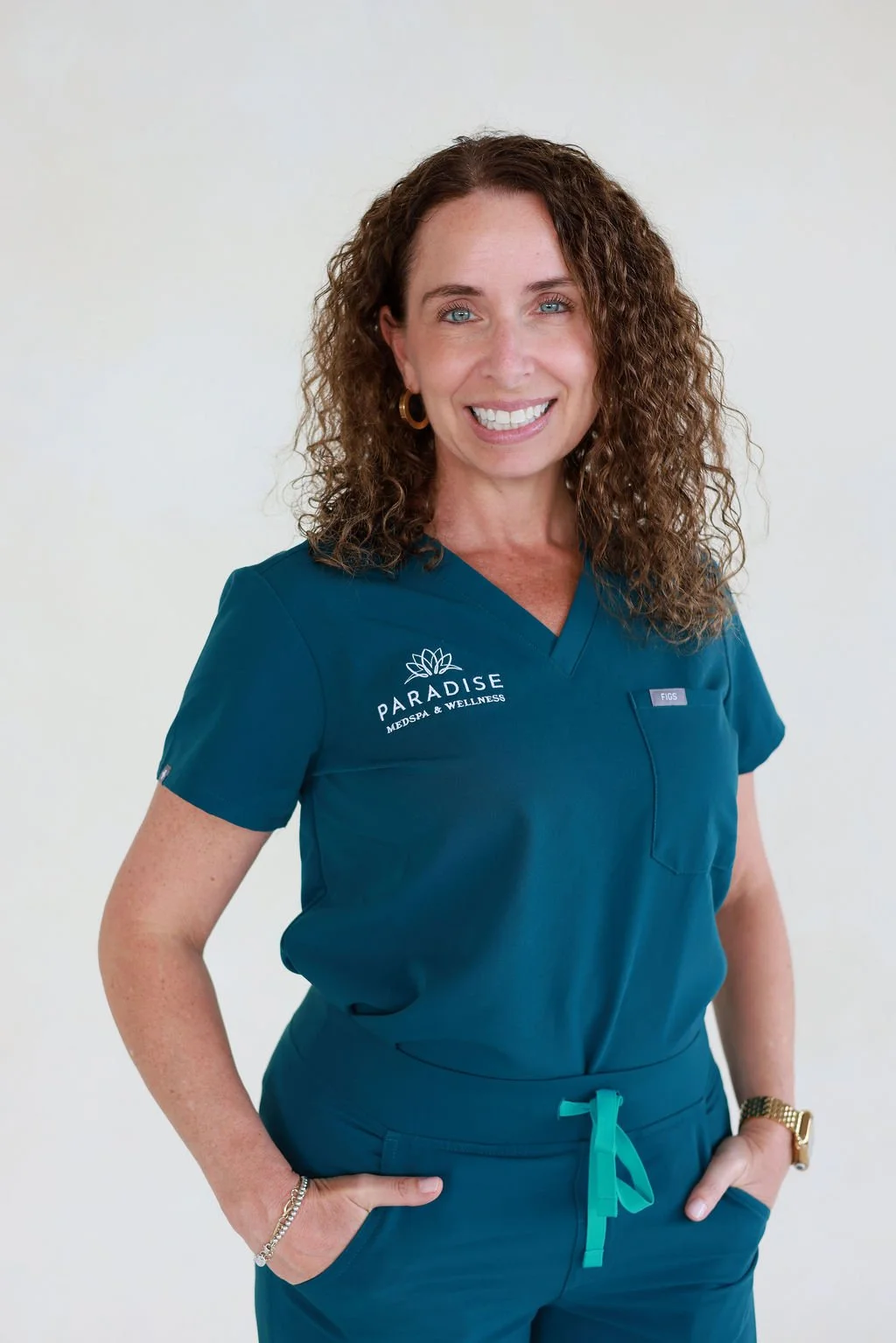 A smiling woman with curly brown hair wearing teal medical scrubs with 'Paradise Medspa & Wellness' logo and name tag.