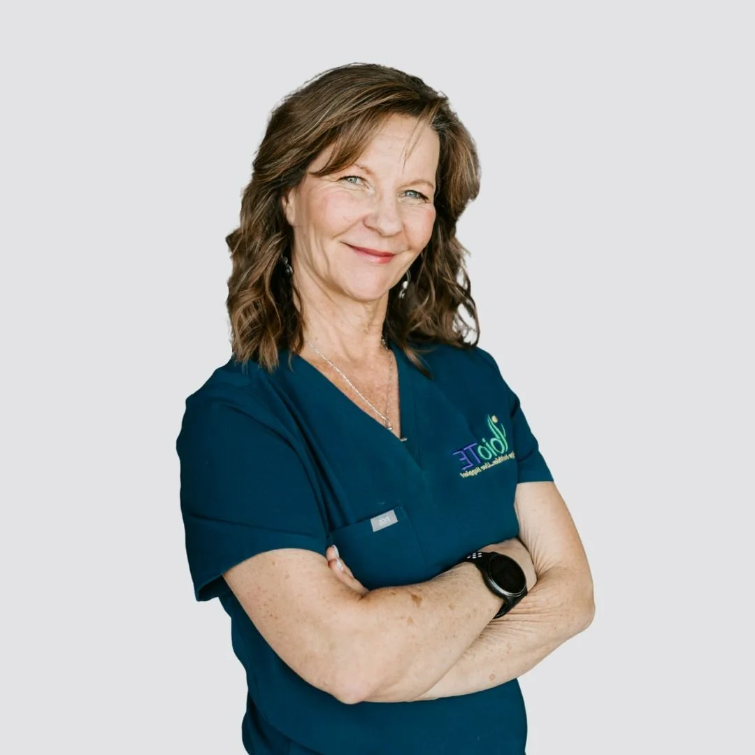 A woman with brown hair, dressed in navy scrubs with a logo, standing with arms crossed and smiling at the camera.
