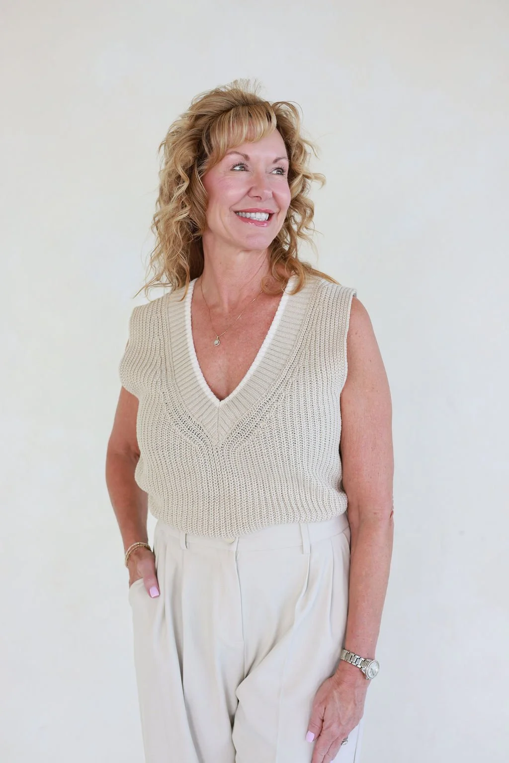A smiling middle-aged woman with blonde, curly hair, wearing a beige sleeveless knit top and beige trousers, standing against a plain off-white background.