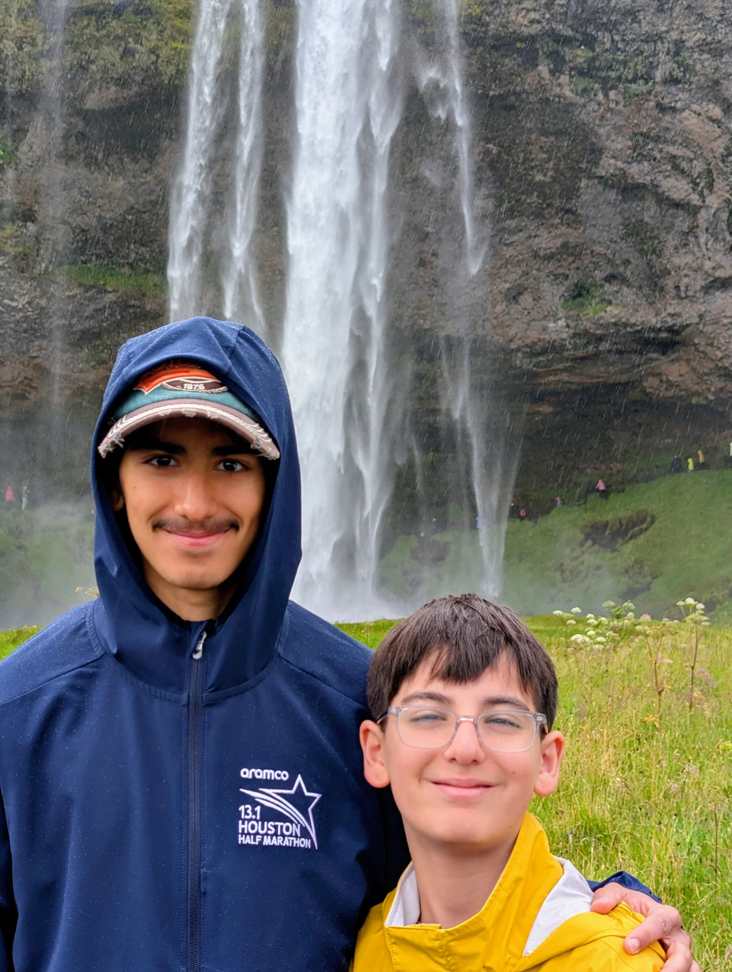 A young man with a beard and a young boy with glasses taking a selfie in front of a waterfall. The young man is wearing a blue hoodie with a logo that reads 'Houston Half Marathon,' and the boy is wearing a yellow jacket. The background features a large waterfall surrounded by greenery and rocks.