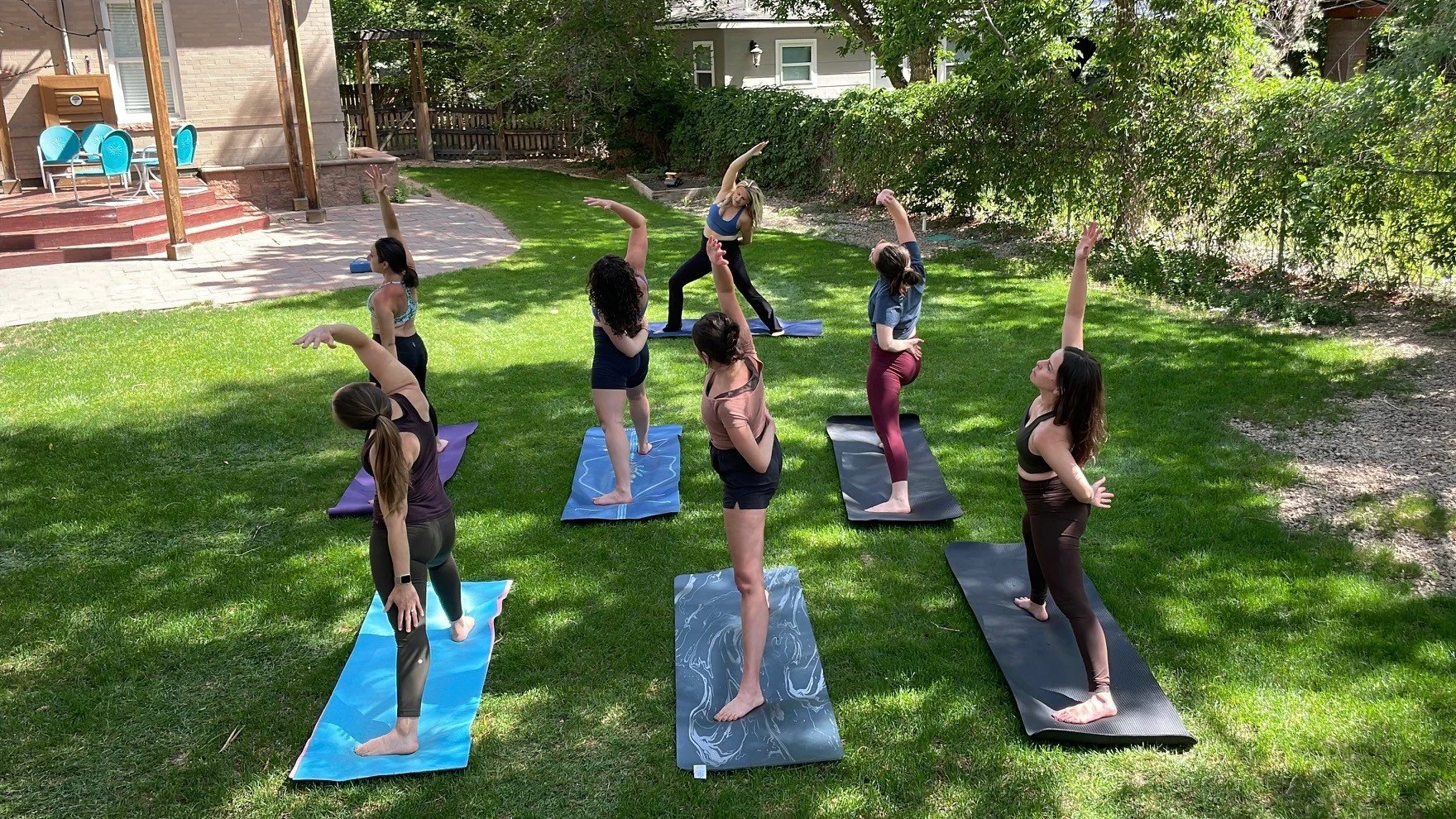 Six women participating in an outdoor yoga class on a grassy backyard, performing a side stretch pose on yoga mats.
