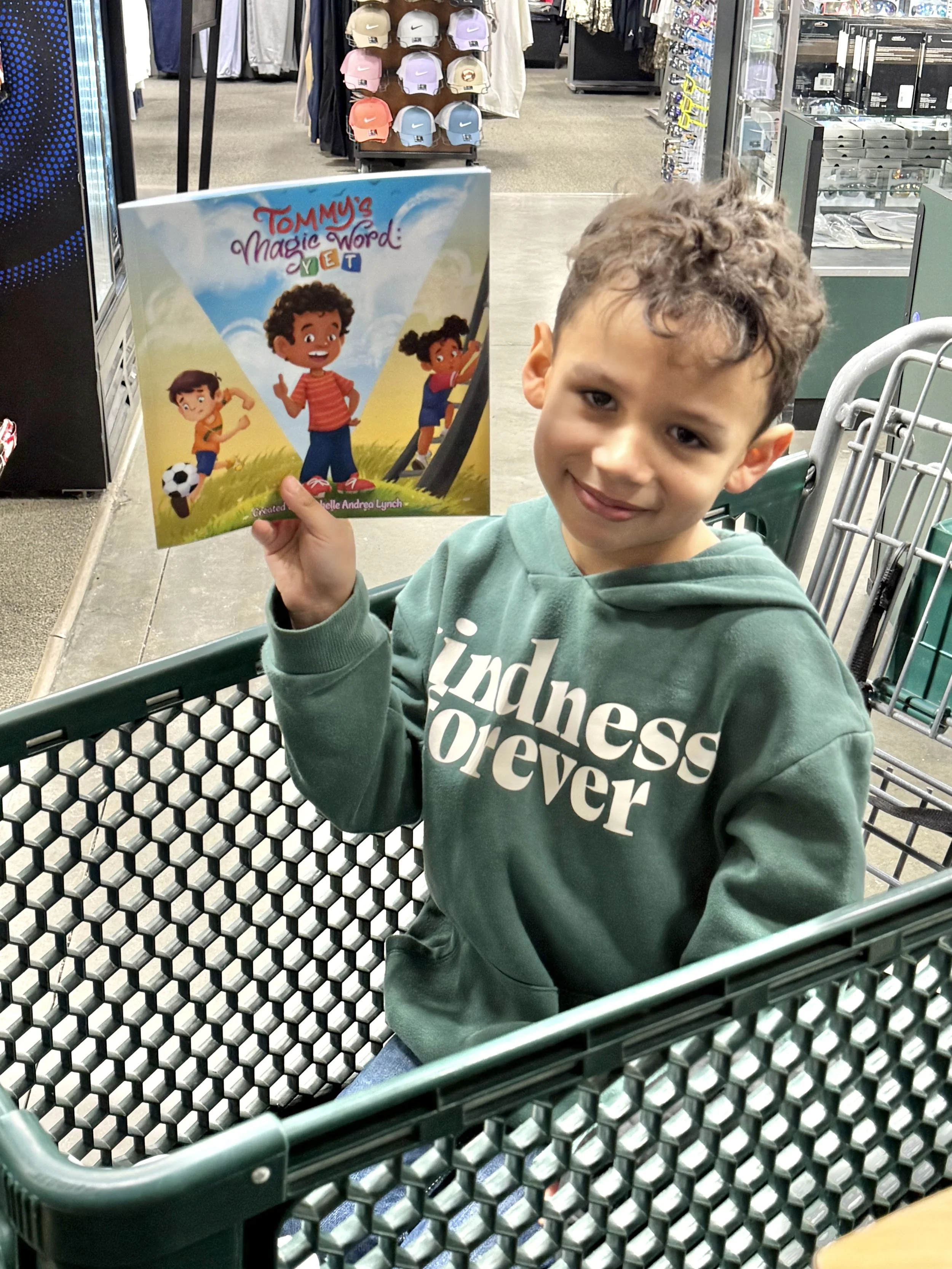A young boy sitting in a shopping cart inside a store, holding up a children's book titled "Tommy's Magic Word: Yet" with colorful illustrations on the cover. The store aisle has clothing and accessories on display in the background.
