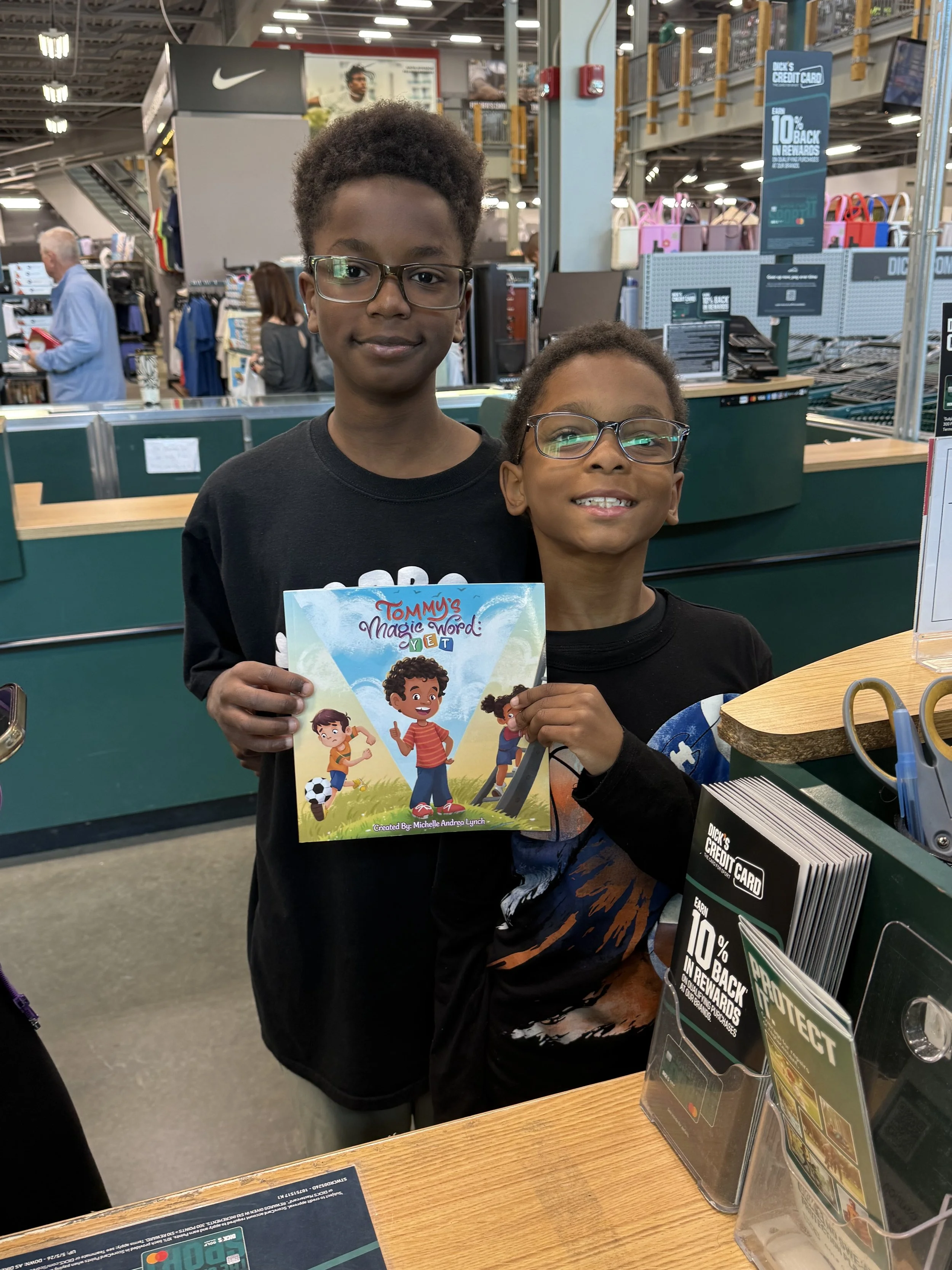 Two children standing at a checkout counter in a store, holding a colorful children's book titled "Tommy's Magic Word: YET." The older child has glasses and is smiling, while the younger child is also smiling and wearing glasses. There are store shel