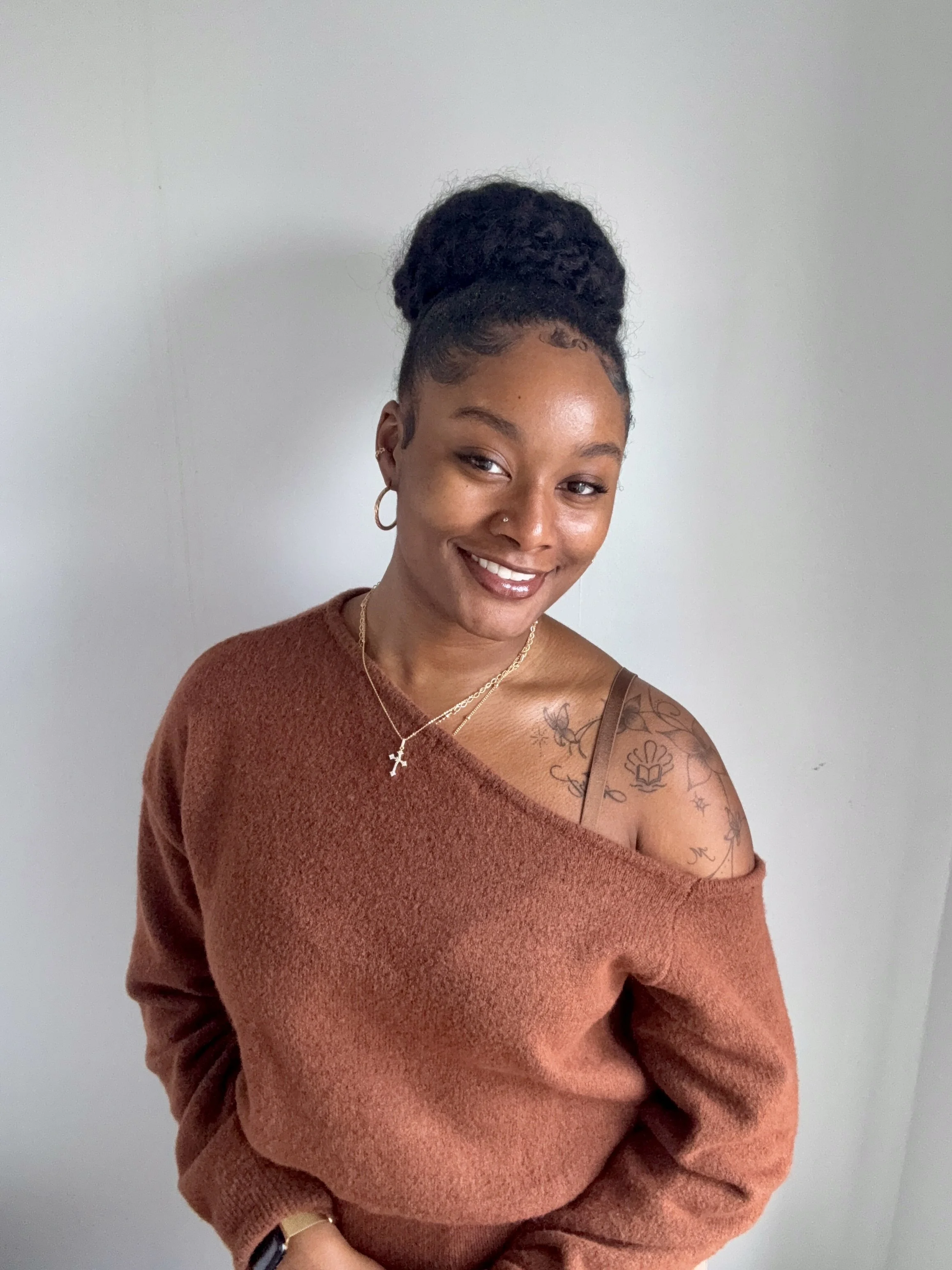 A young woman smiling with her eyes slightly closed, wearing a brown off-shoulder sweater, gold jewelry including earrings, a necklace with a cross, and a watch, standing against a plain off-white wall. She has dark hair styled in a high bun and visible tattoos on her shoulder.