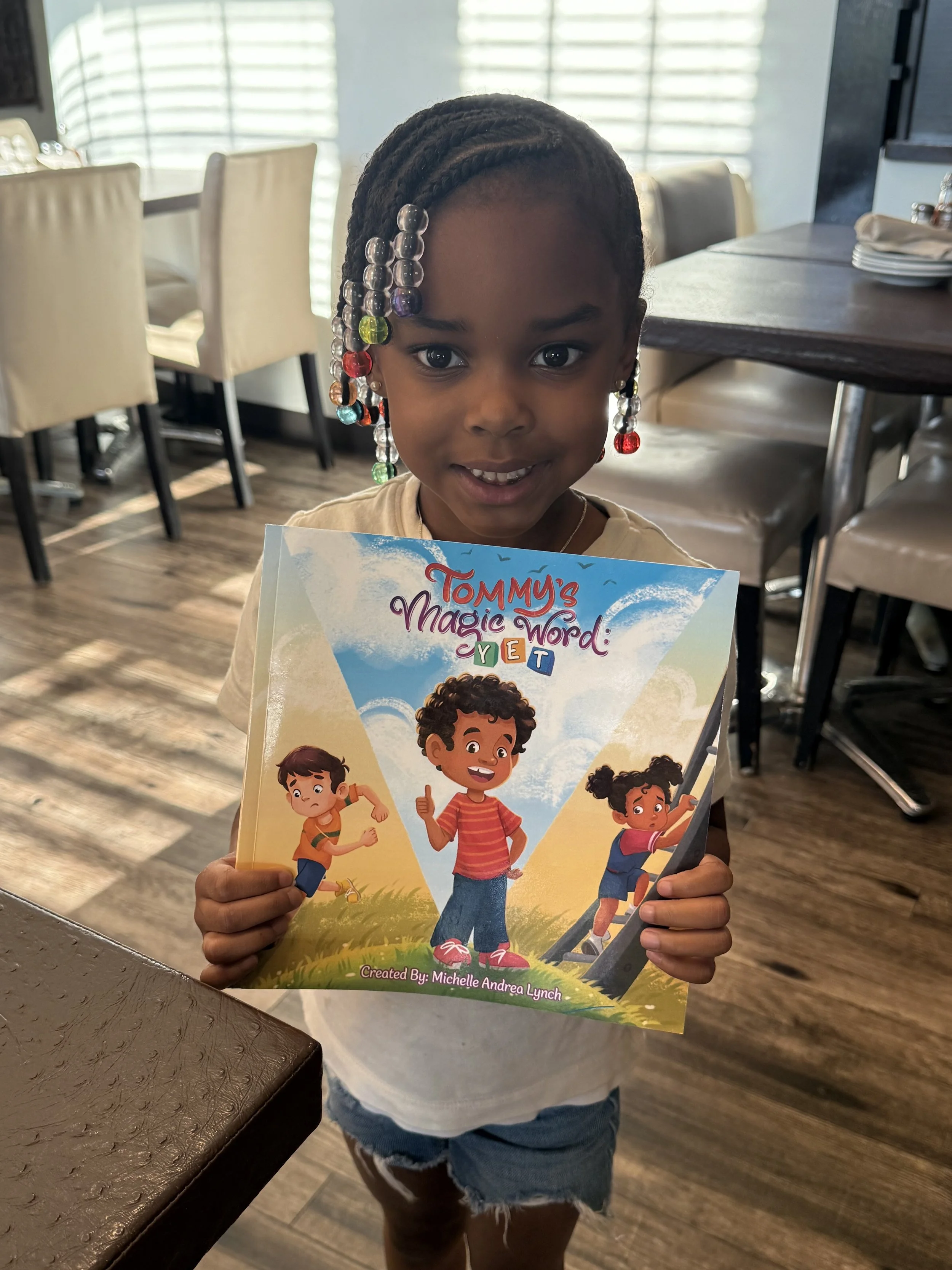 A young girl with braided hair decorated with colorful beads is holding a children's book titled 'Tommy's Magic Word' in a restaurant setting. She is smiling and standing near a table with stacked plates and napkins. The background features chairs an