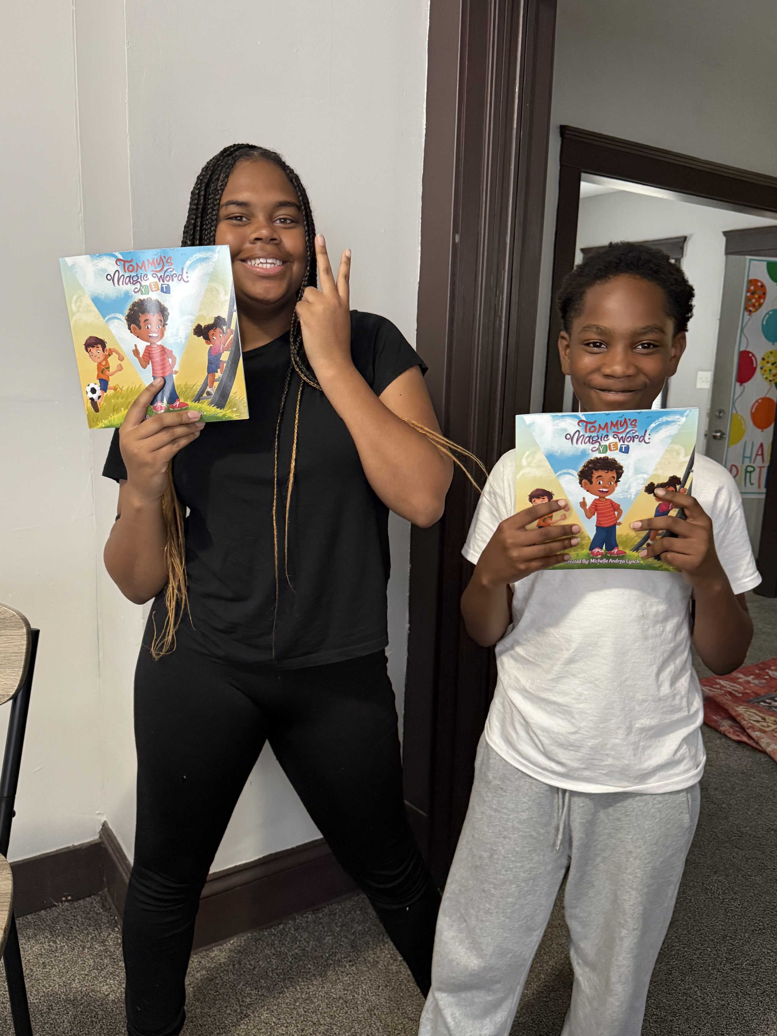 Two children standing indoors, holding books titled "Tommy's Magic Word". The girl on the left is smiling and making a peace sign with her fingers. The boy on the right is smiling and looking at the camera.