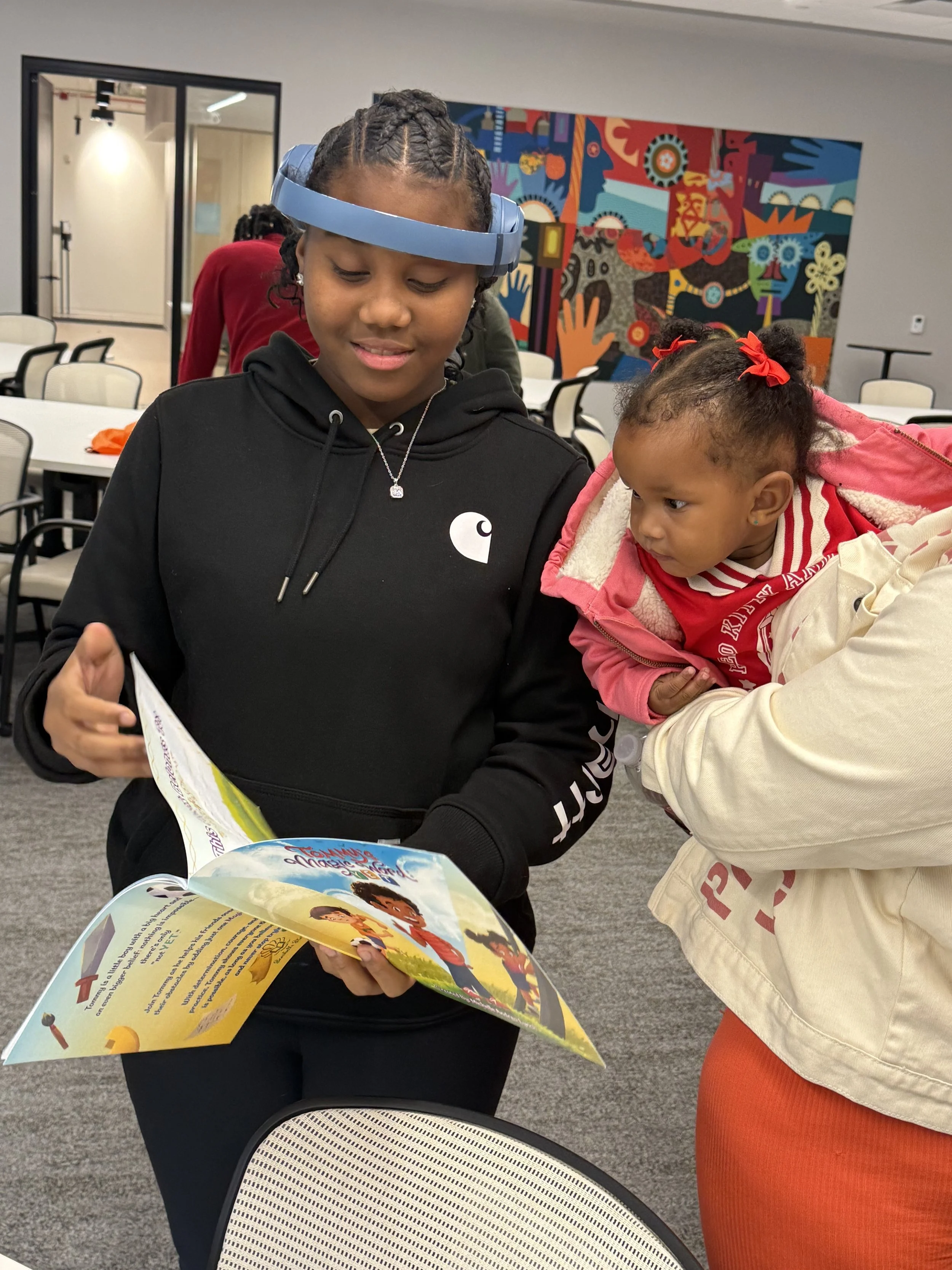 A woman with braided hair and a light blue headband is holding a colorful children's book named 'Tommy's Magic Word: Yet', showing it to a young girl wearing red bows in her hair, in a room with a vibrant mural on the wall.