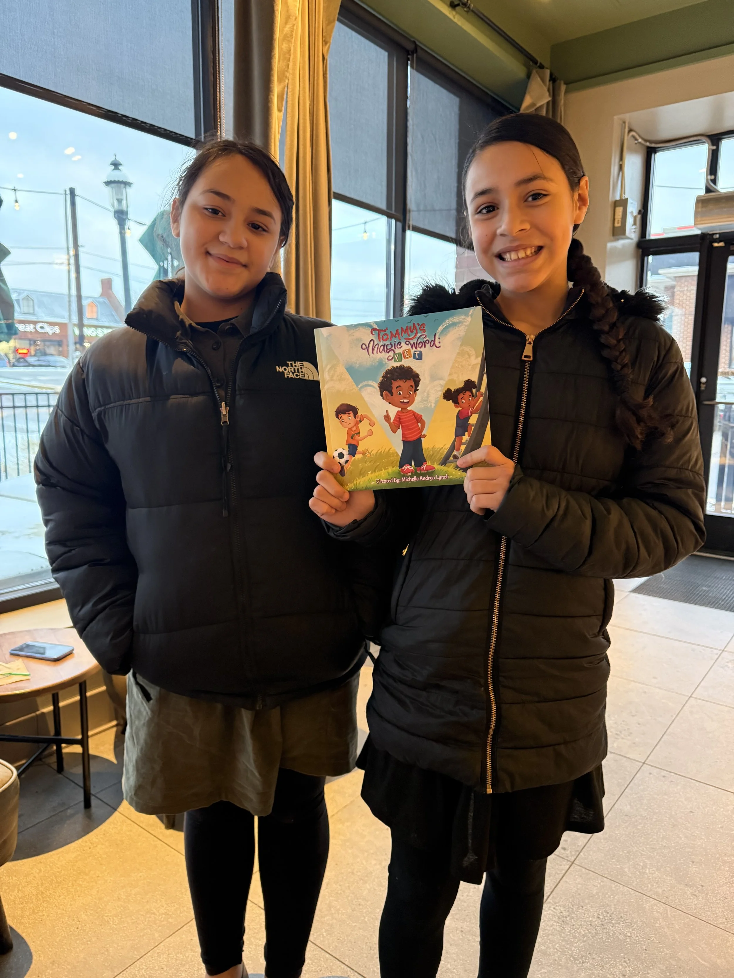 Two girls with dark hair in braids, wearing black jackets, holding a colorful book titled 'Tommy's Magic Word: Yet' in a restaurant or cafe with large windows and yellow curtains.