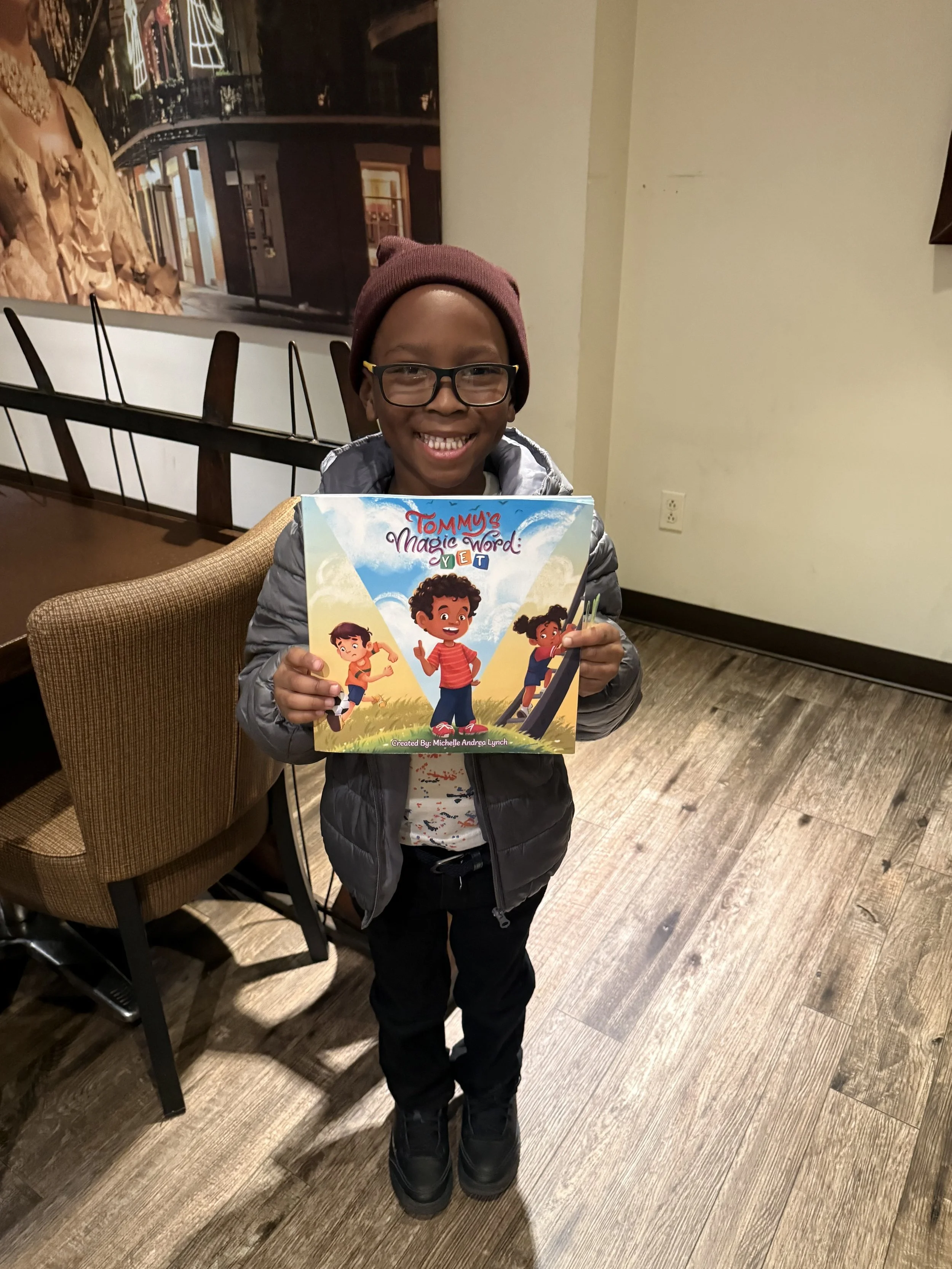 A smiling young boy wearing glasses, a maroon beanie, a gray jacket, and black pants, holding a colorful book titled 'Tommy's Magic Word: YET' in a room with wooden flooring and a beige wall.