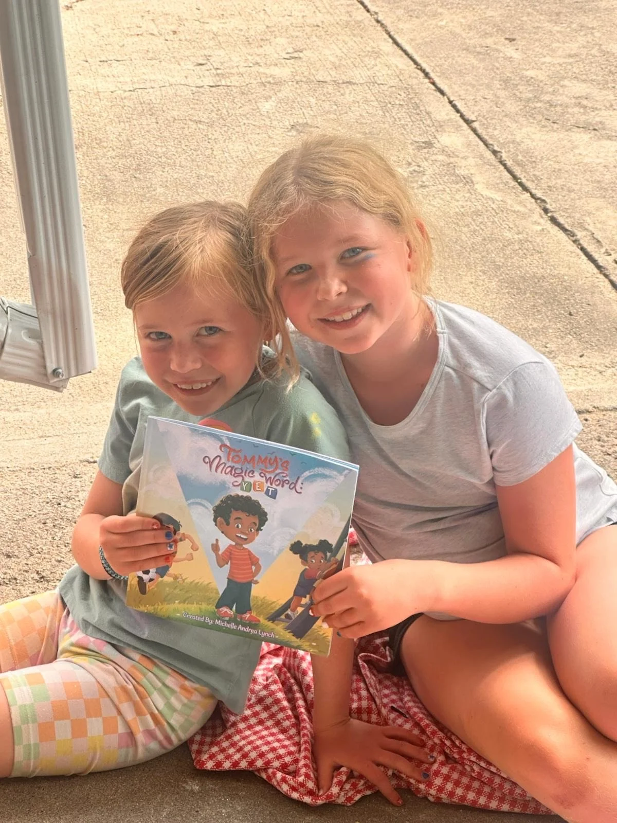 Two young girls sitting on a concrete surface, smiling at the camera. The younger girl holds a colorful book titled "Tommy's Magic Word: Yet." The background shows a concrete sidewalk and the metal pole of an outdoor structure.