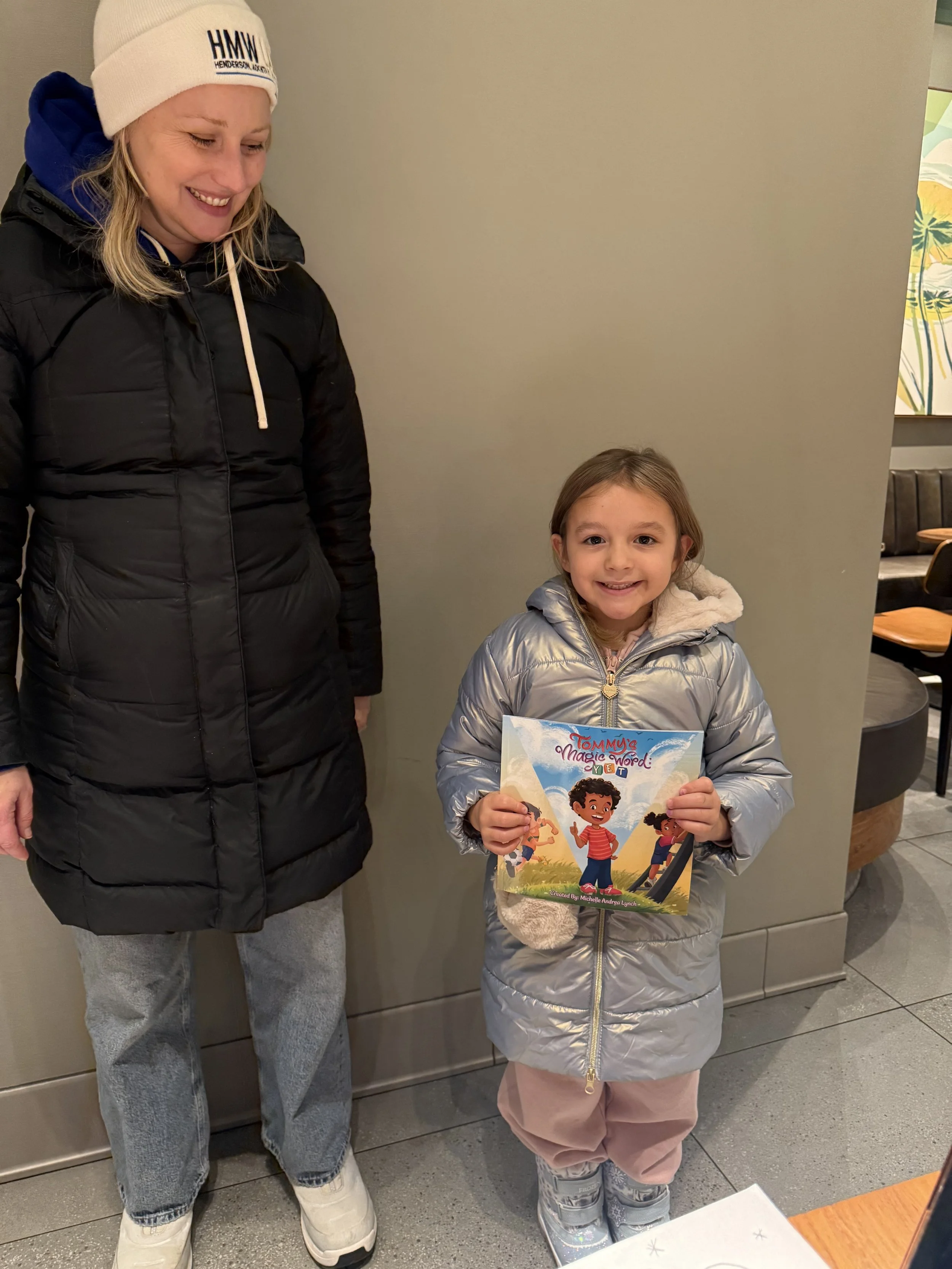 A young girl in a silver puffer jacket holding a colorful children's book titled 'Tommy's Magic Word: Yet' while smiling at the camera, standing next to a woman in a black winter coat and white hat, inside a cafe or restaurant.