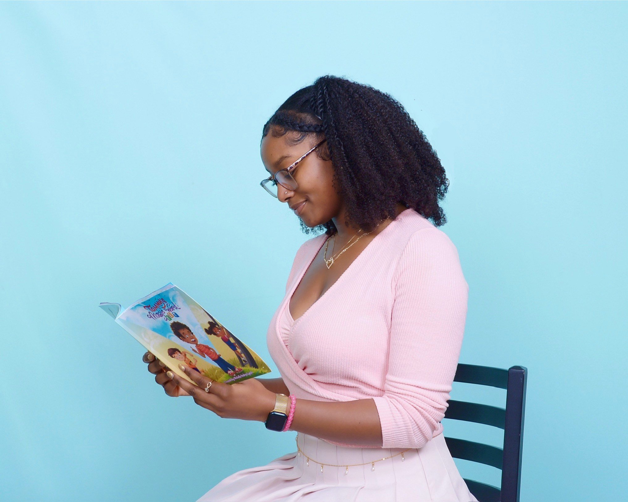 A woman with curly hair, glasses, and jewelry, wearing a pink sweater, is sitting on a blue chair against a light blue background, reading a colorful children's book.