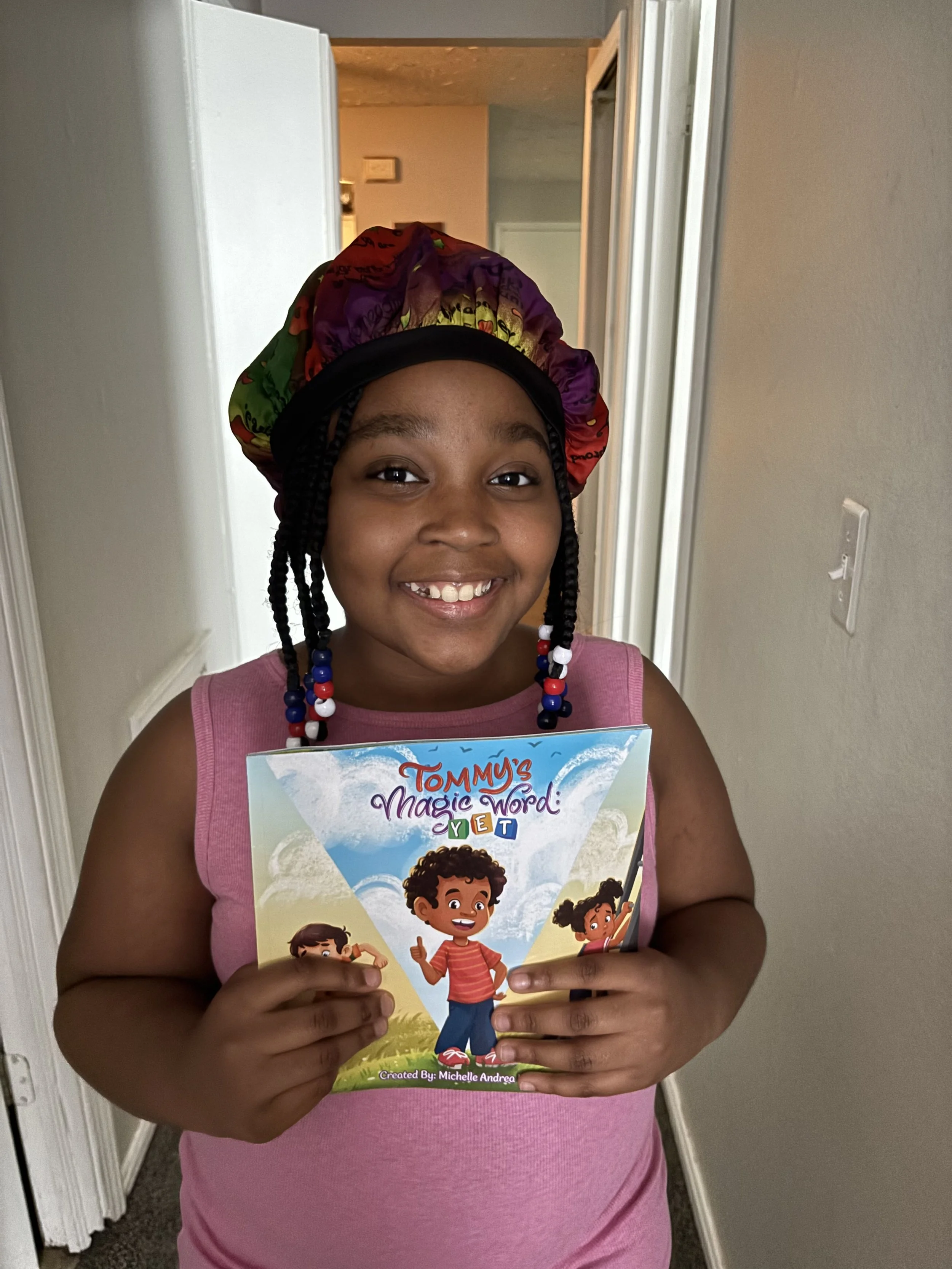A young girl with braids and a colorful bonnet holding a children's book titled "Tommy's Magic Word: Yet" and smiling at the camera.