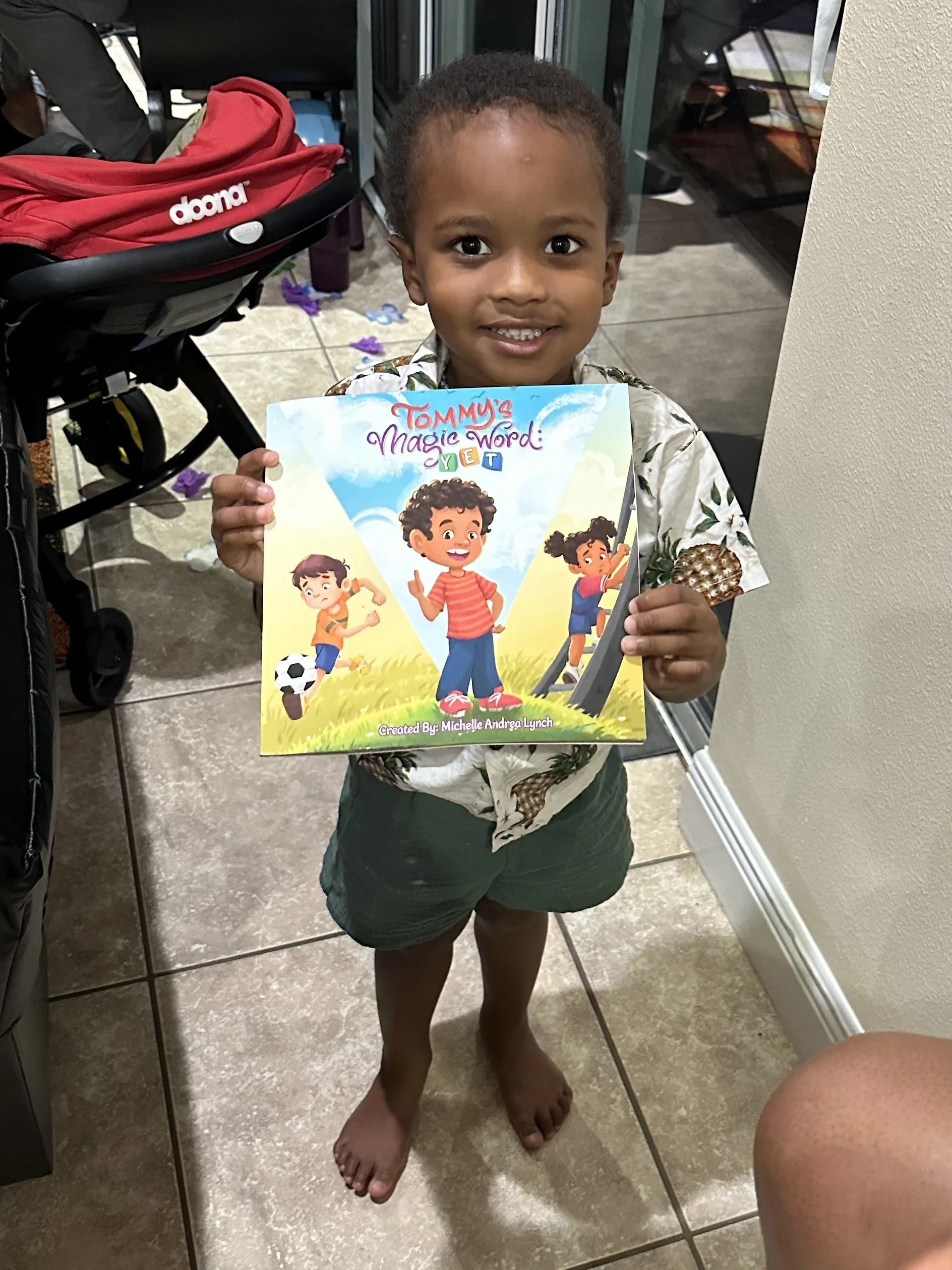 A young boy with dark curly hair smiling and holding a children's book titled "Tommy's Magic Word: YET" in an indoor setting with a stroller and tiled floor.