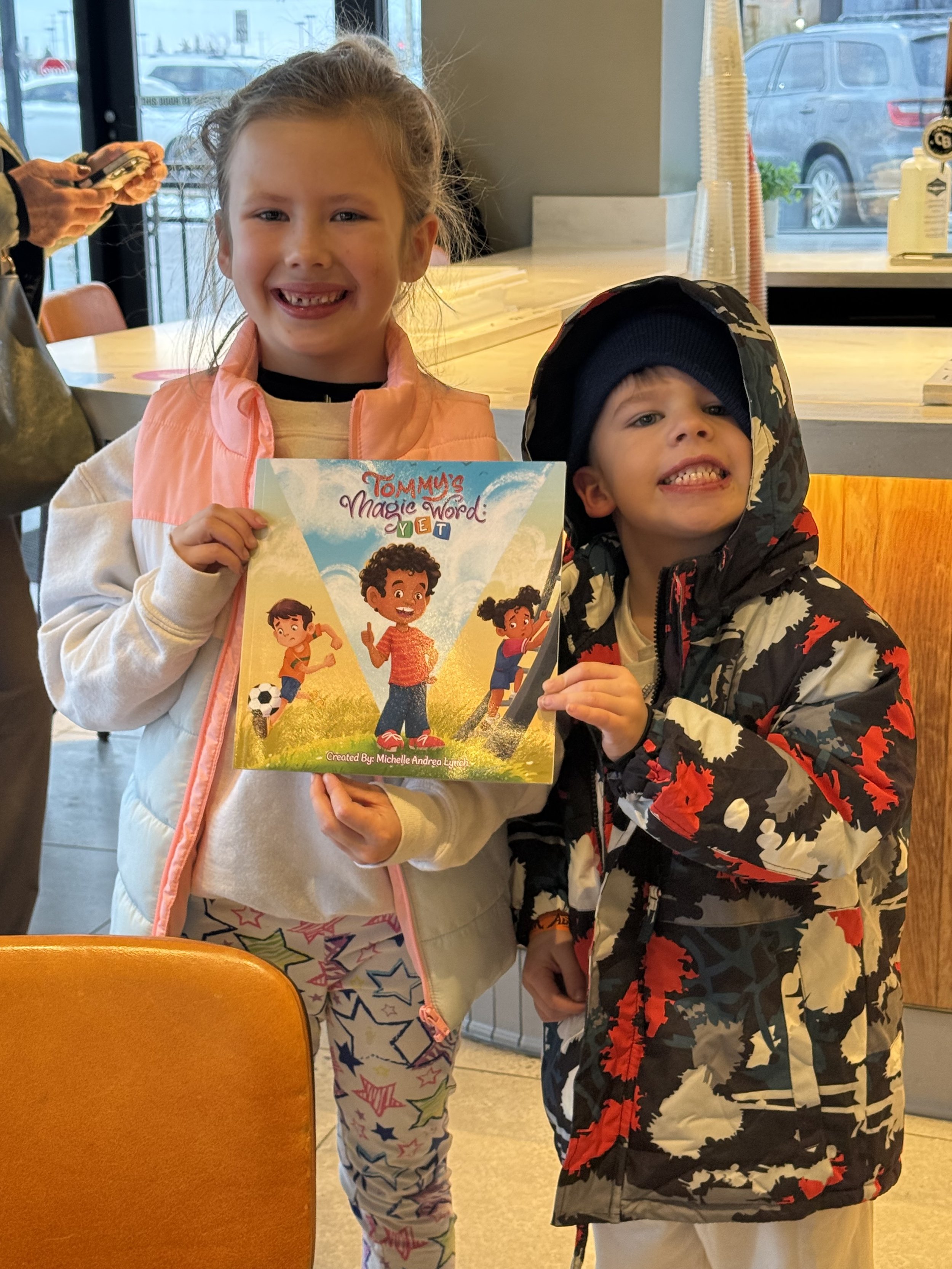 Two children, a girl and a boy, posing inside a cafe. The girl holds a colorful book titled 'Tommy's Magic Word YET.' The girl has curly hair, is smiling, and wears a light-colored jacket with a pink front. The boy is wearing a black beanie, a camouf