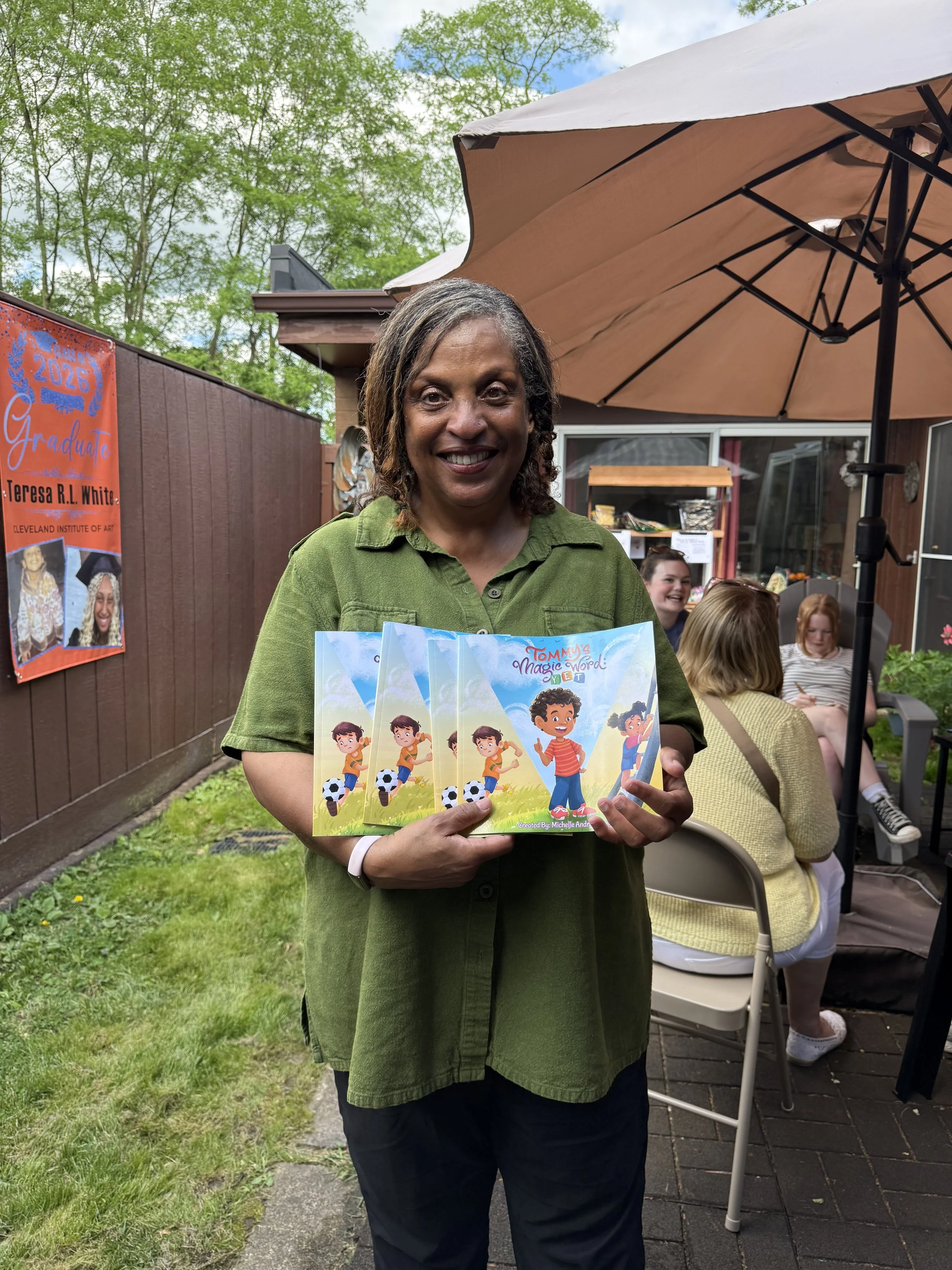 Woman smiling and holding children’s books titled 'Tommy's Magic Word: Yet' at an outdoor gathering, surrounded by trees, a brown fence, and other people sitting and talking under a large tan umbrella.