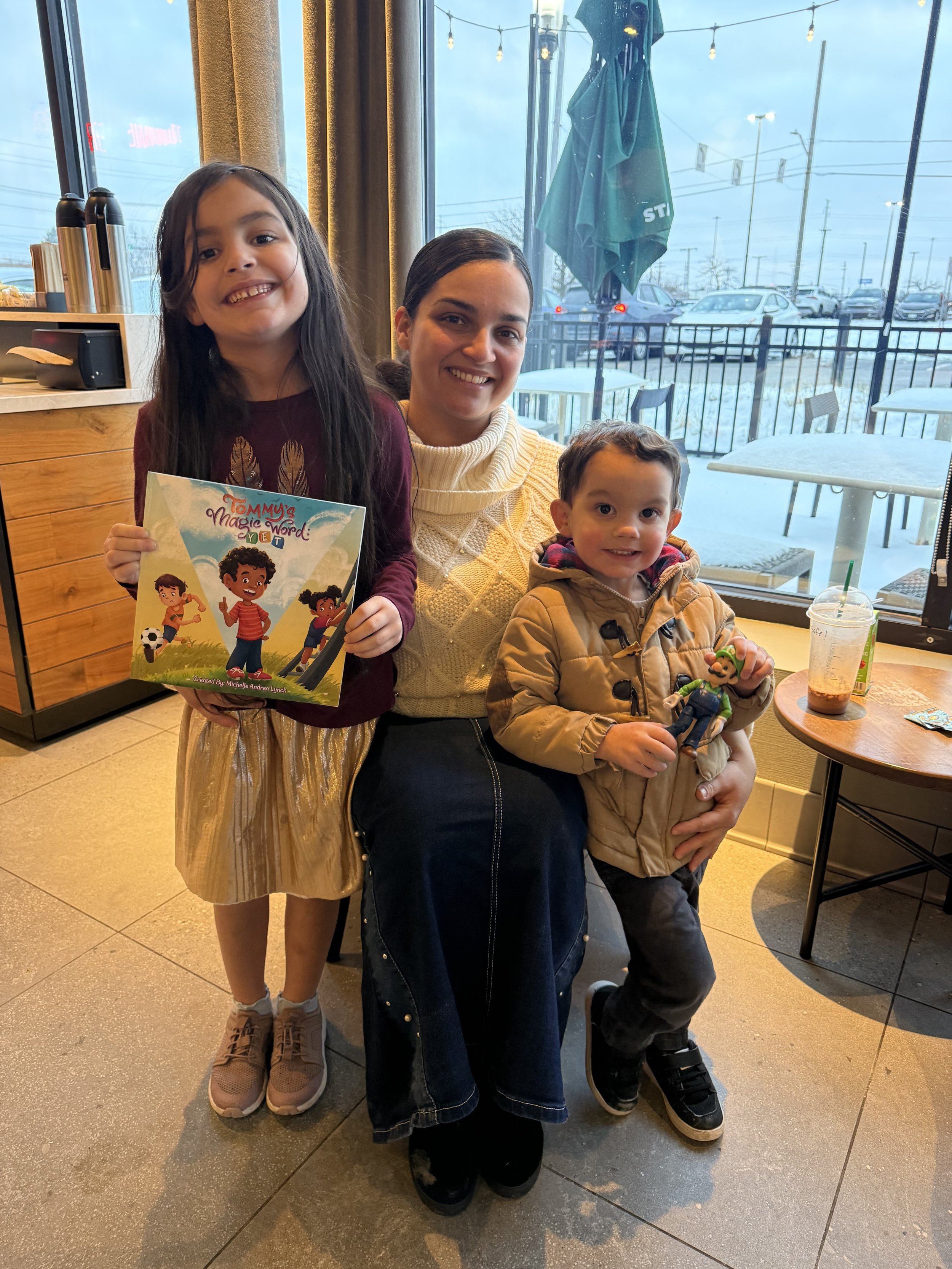 Three children smiling inside a cafe, with large windows showing a snowy parking lot and street outside. The girl on the left is holding a colorful storybook, and the boy on the right is holding a small plush toy. They are standing close together, dr