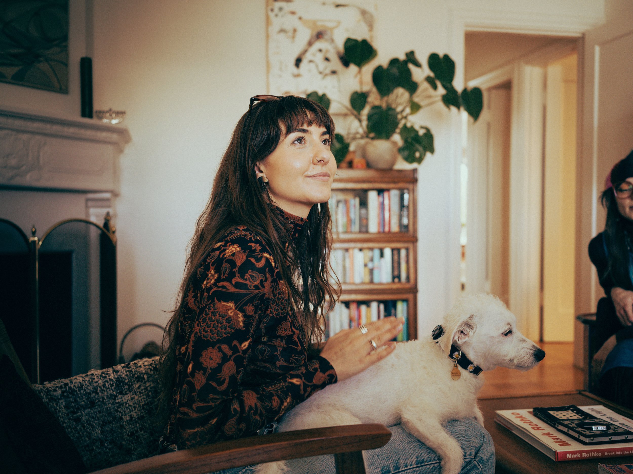 A woman with dark brown hair and a patterned dress is sitting on a couch, petting a large white dog with a collar. She is smiling and looking slightly upward. In the background, there is a bookshelf with books, a large potted plant, and a wall with hanging art. To her right, a partially visible girl with glasses is sitting, and a table in front of her has some books