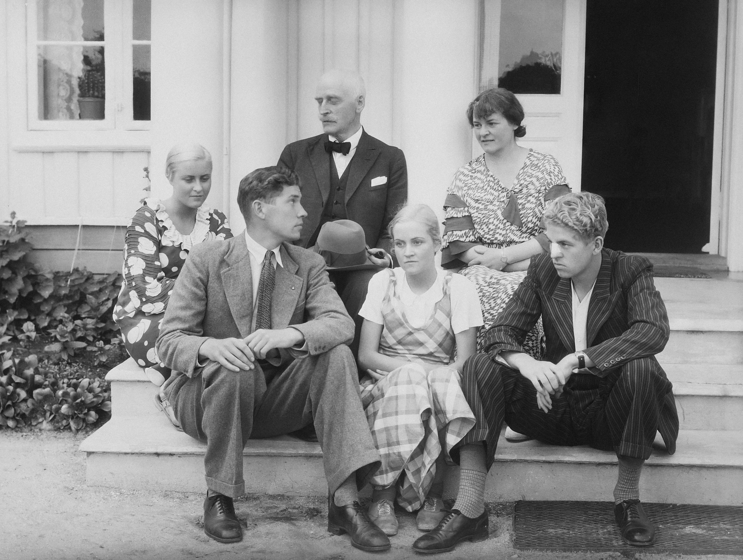 A black and white photo of six young people and one older man sitting and standing on a porch in front of a house. The man and some of the girls are dressed in vintage clothing from the 1940s or 1950s.