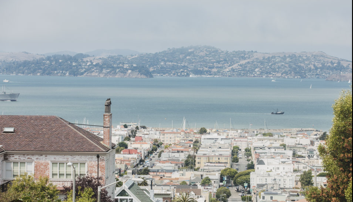 Overlooking a coastal city with a bay and hilly landscape in the background, and buildings, streets, and trees in the foreground.