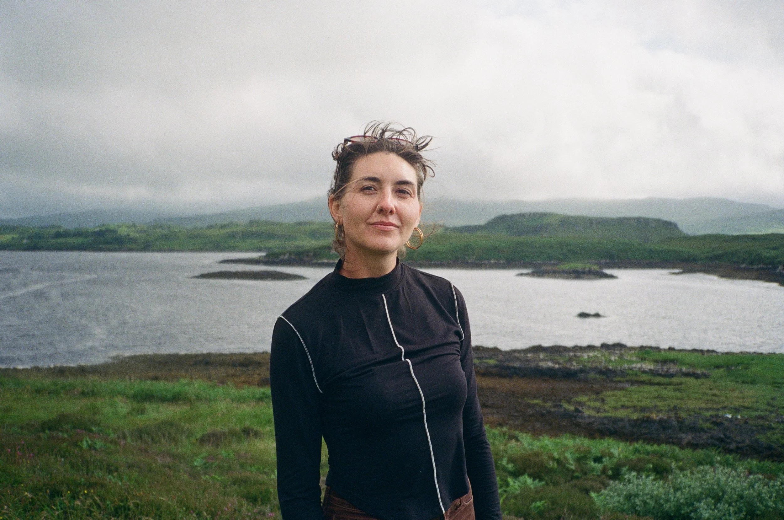 A young woman with wavy brown hair, earrings, and a dark long-sleeve top stands outdoors near a body of water, with grassy land and green hills in the background under a cloudy sky.